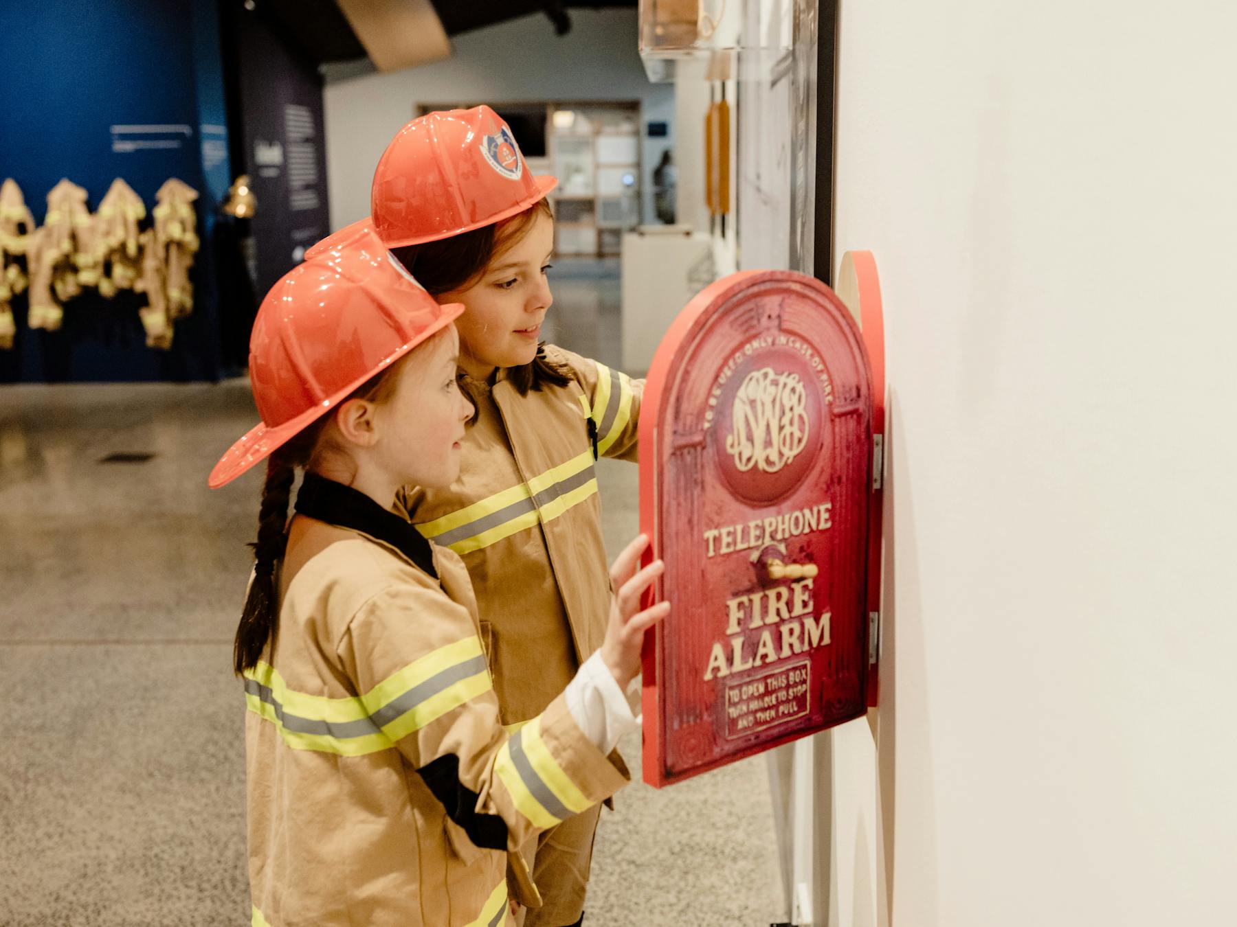 Two children dressed in fire fighter uniforms and hats looking at a replica fire alarm telephone