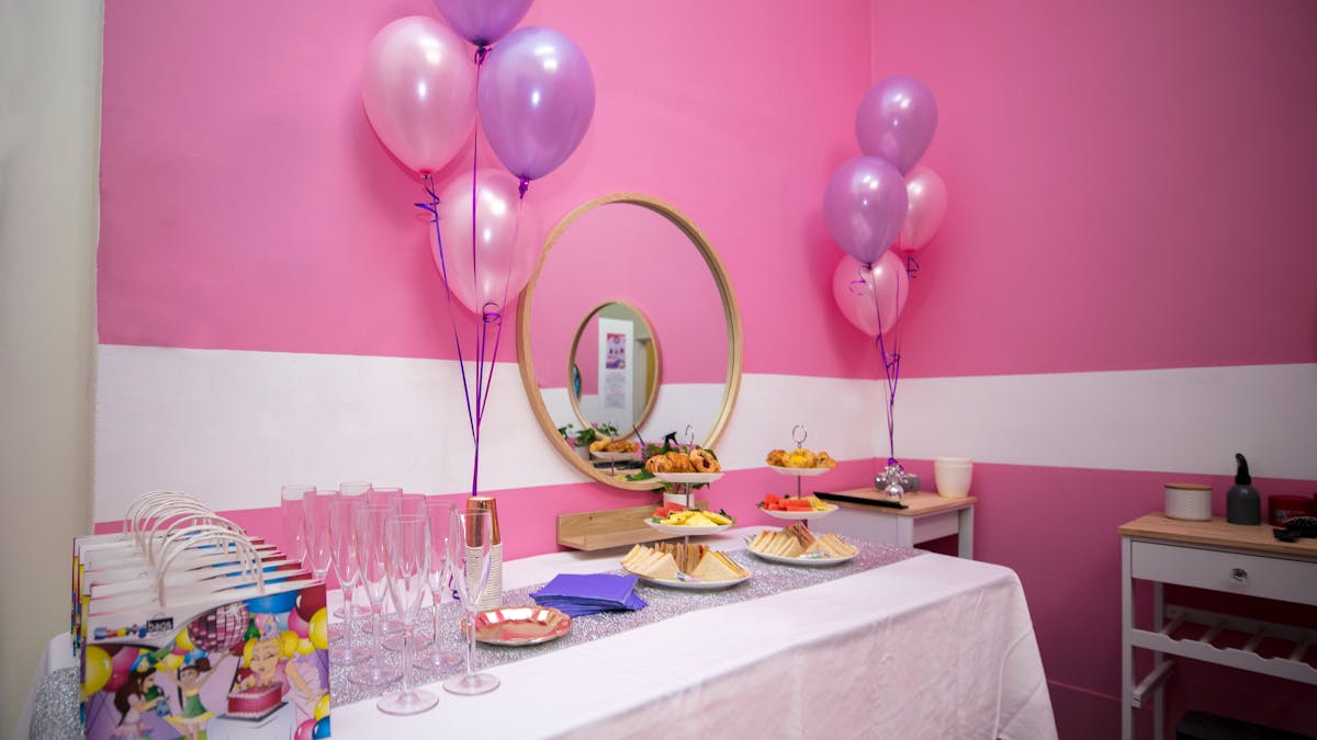 A long table with a white table cloth with pink and purple balloons and party bags