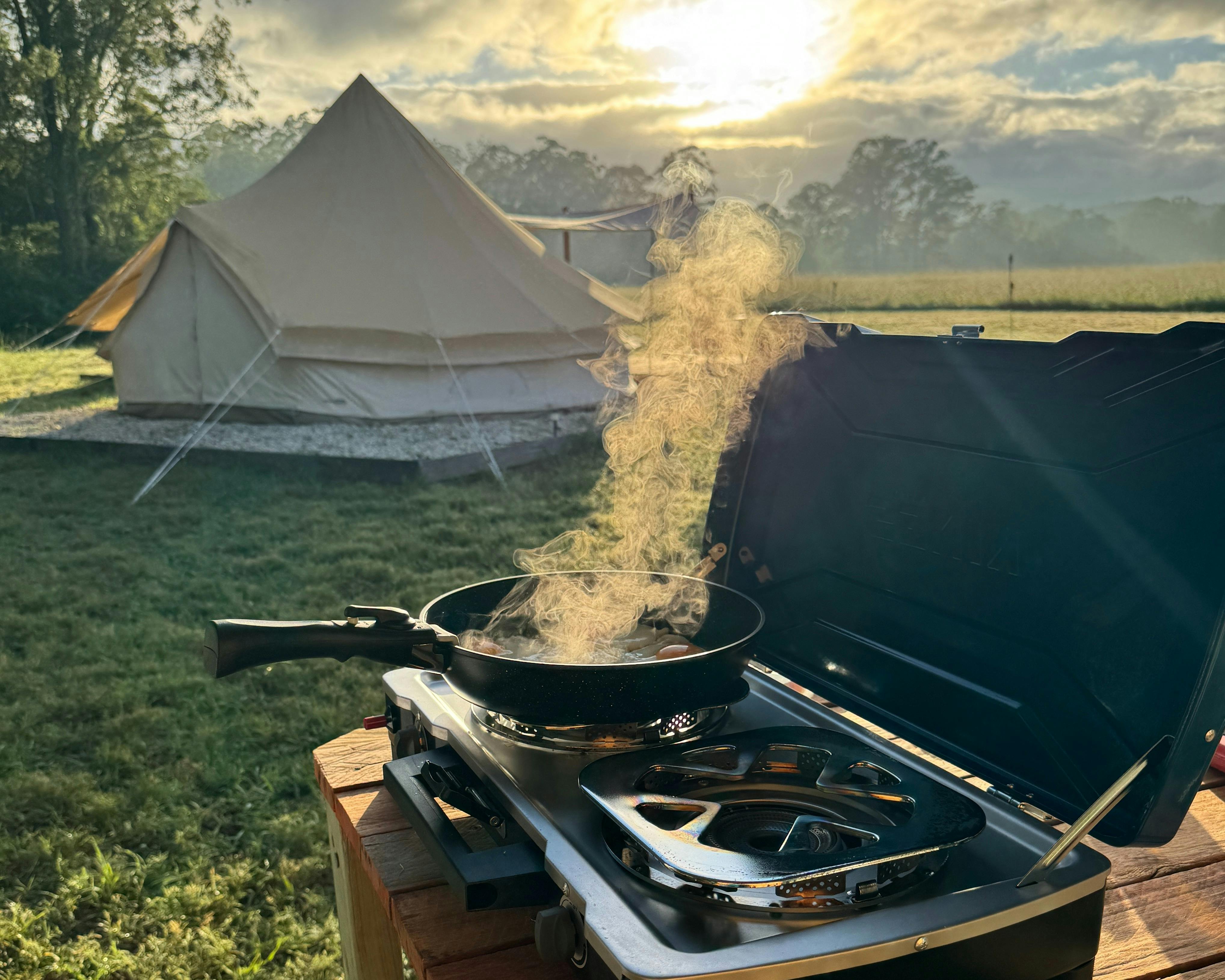 목초지와 텐트 위로 떠오르는 일출을 바라보며 더블 가스 버너 위에서 프라이팬으로 계란을 굽습니다.