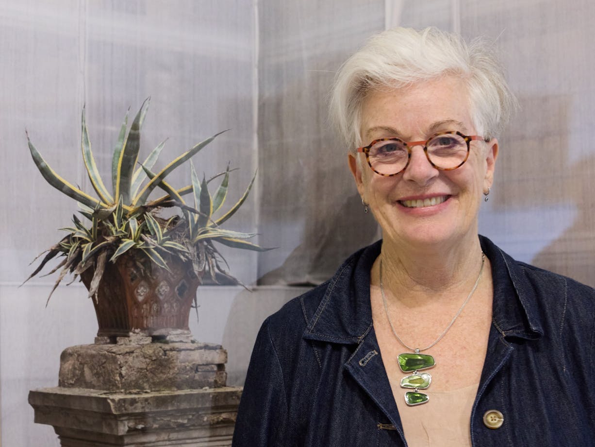 Hilary Wardhaugh smiling, stands in front of a large photograph of a fern growing in a ruined plinth