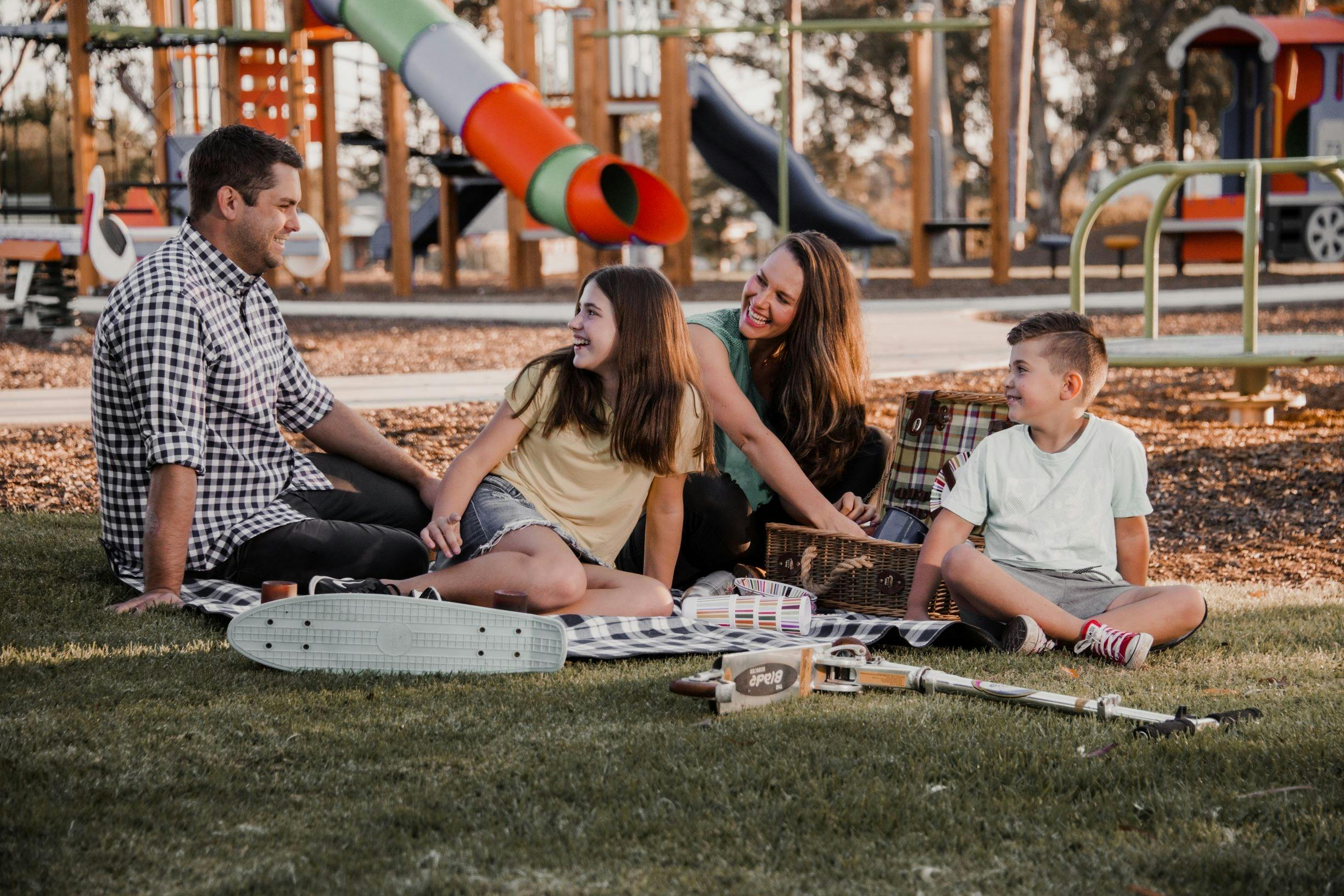 Young family sitting at a lushious green park in front of an amazing playground enjoying the sun