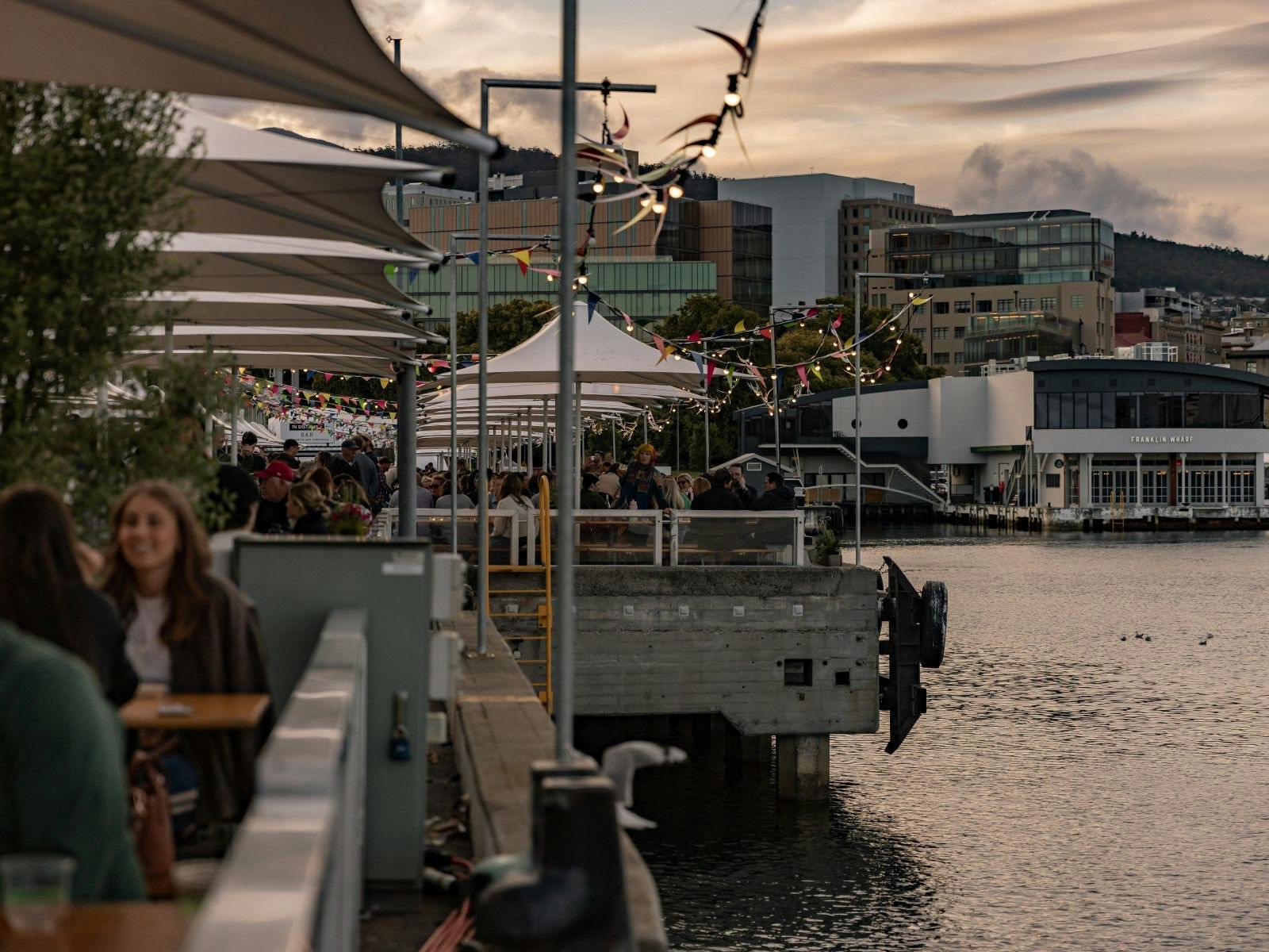 People relax by Hobart's pier at sunset beneath bunting and soft lights near still waters.