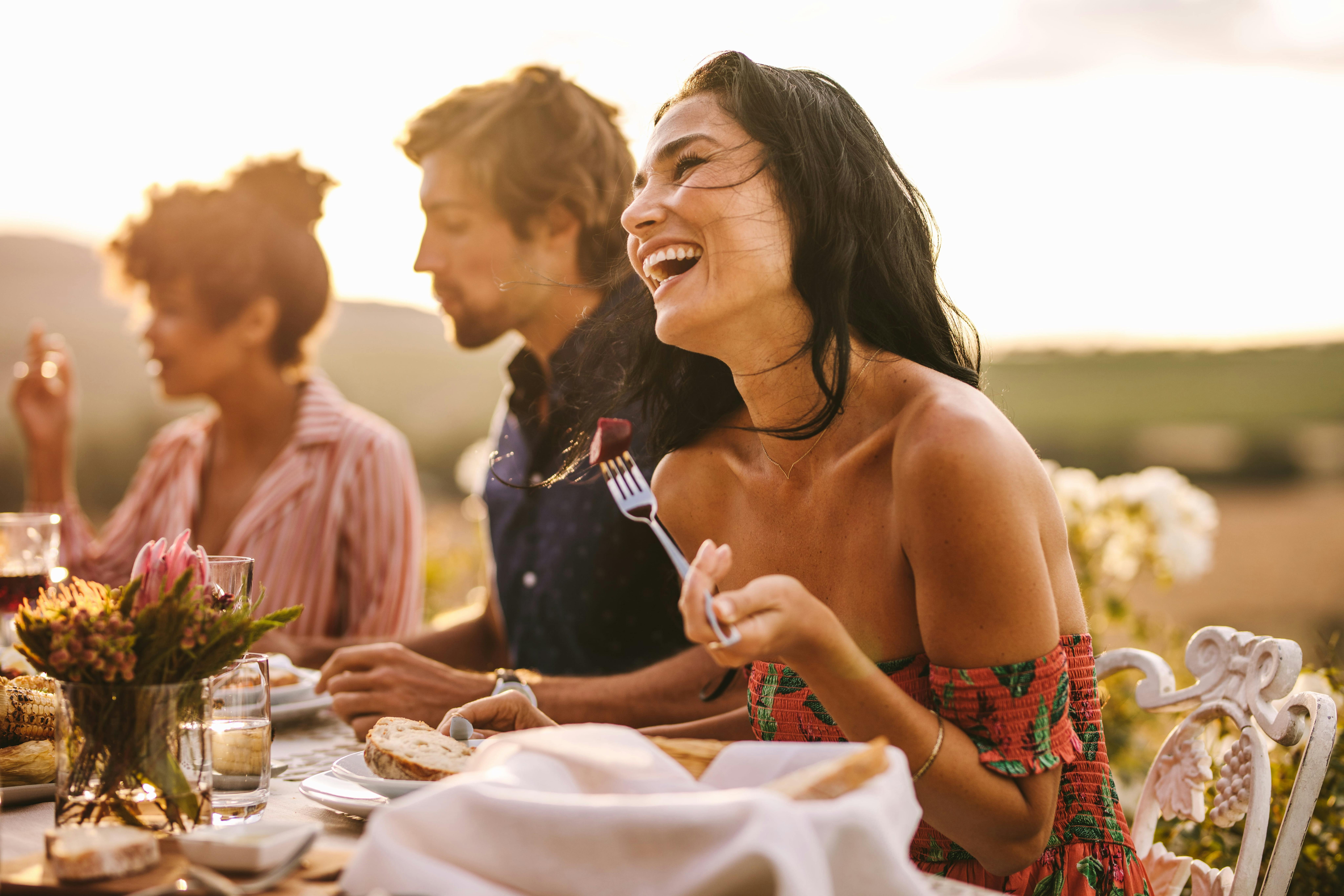 Dark haired woman in the foreground laughing with a fork in her hand. A male and female in the back
