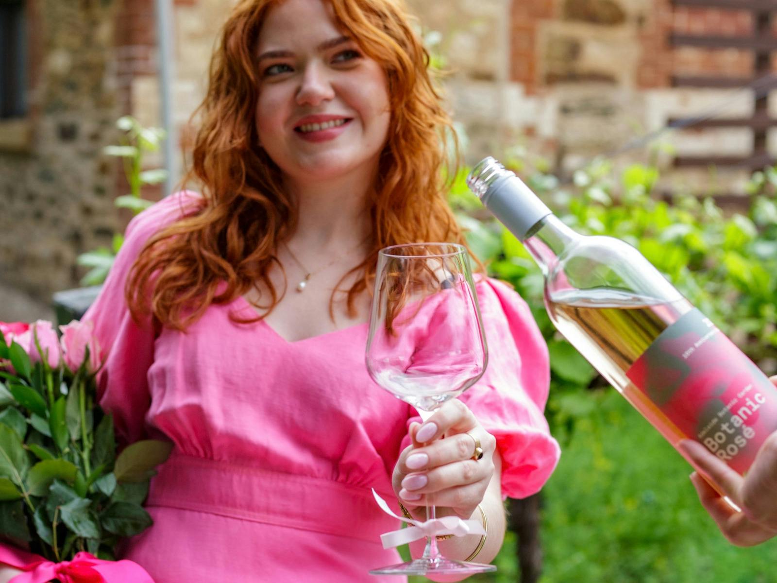 Smiling red-haired woman in a bright pink dress holds a wineglass beside an angled bottle