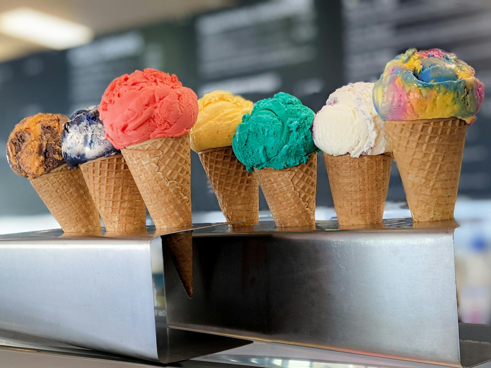 Seven single-scoop ice cream cones, of all different colours and flavours