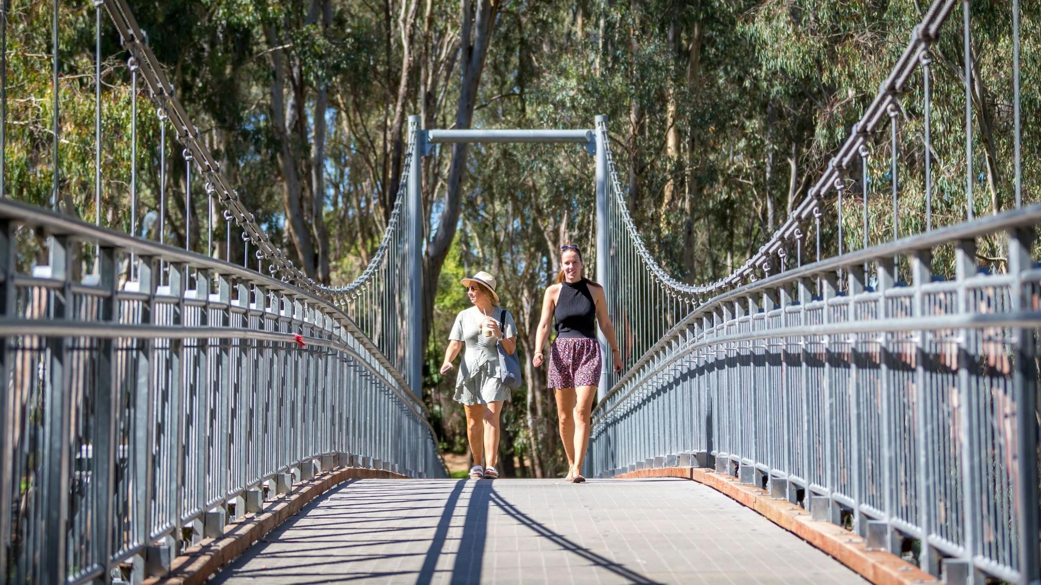 Two walkers walking across swing bridge, trees, sunny day.