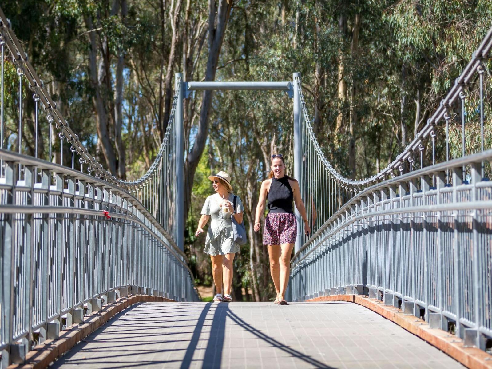 Two walkers walking across swing bridge, trees, sunny day.
