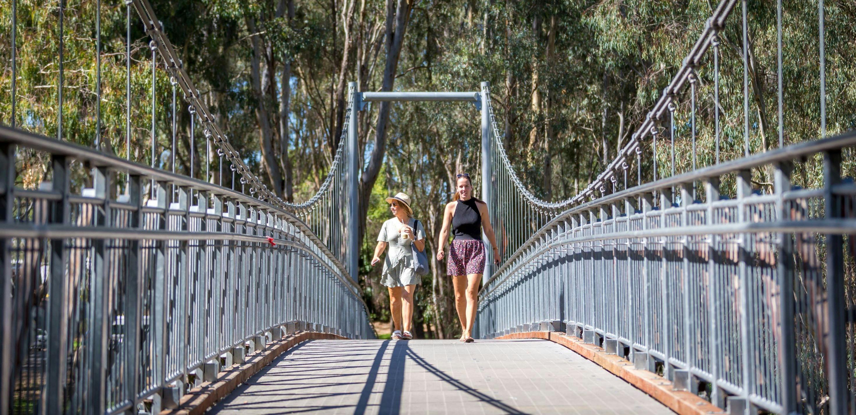 Two walkers walking across swing bridge, trees, sunny day.