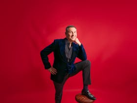 David Walliams behind a red background with his leg on a stool leaning on his knee smiling in a suit