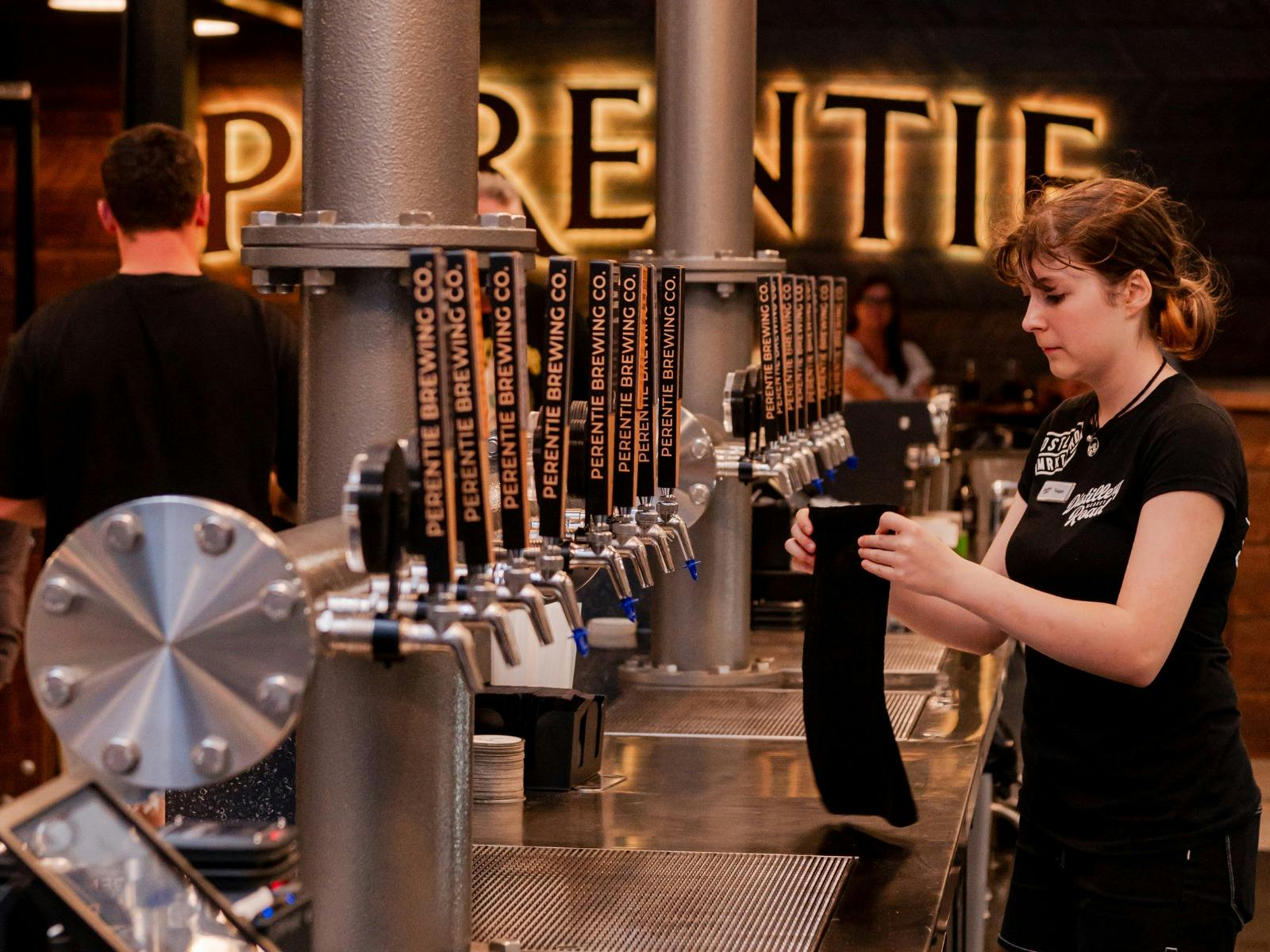 Young female stands behind bar where a row of beer taps can be seen in front of a Perentie sign Young female stands behind bar where a row of beer taps can be seen in front of a Perentie sign