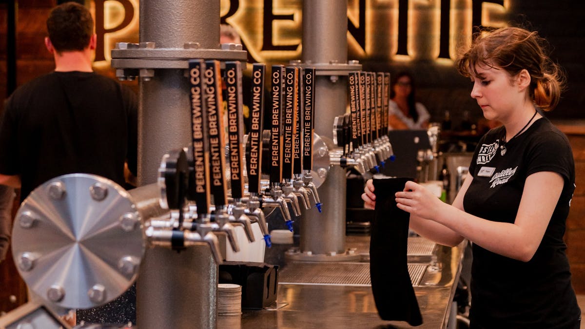 Young female stands behind bar where a row of beer taps can be seen in front of a Perentie sign