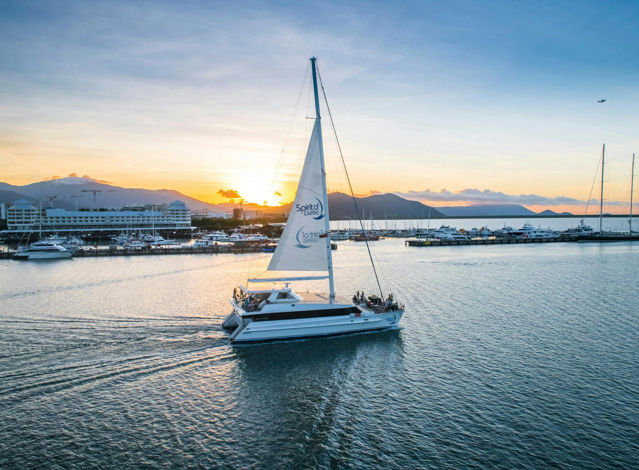 Catamaran sailing down Trinity Inlet with sun setting over mountains and cairns marina