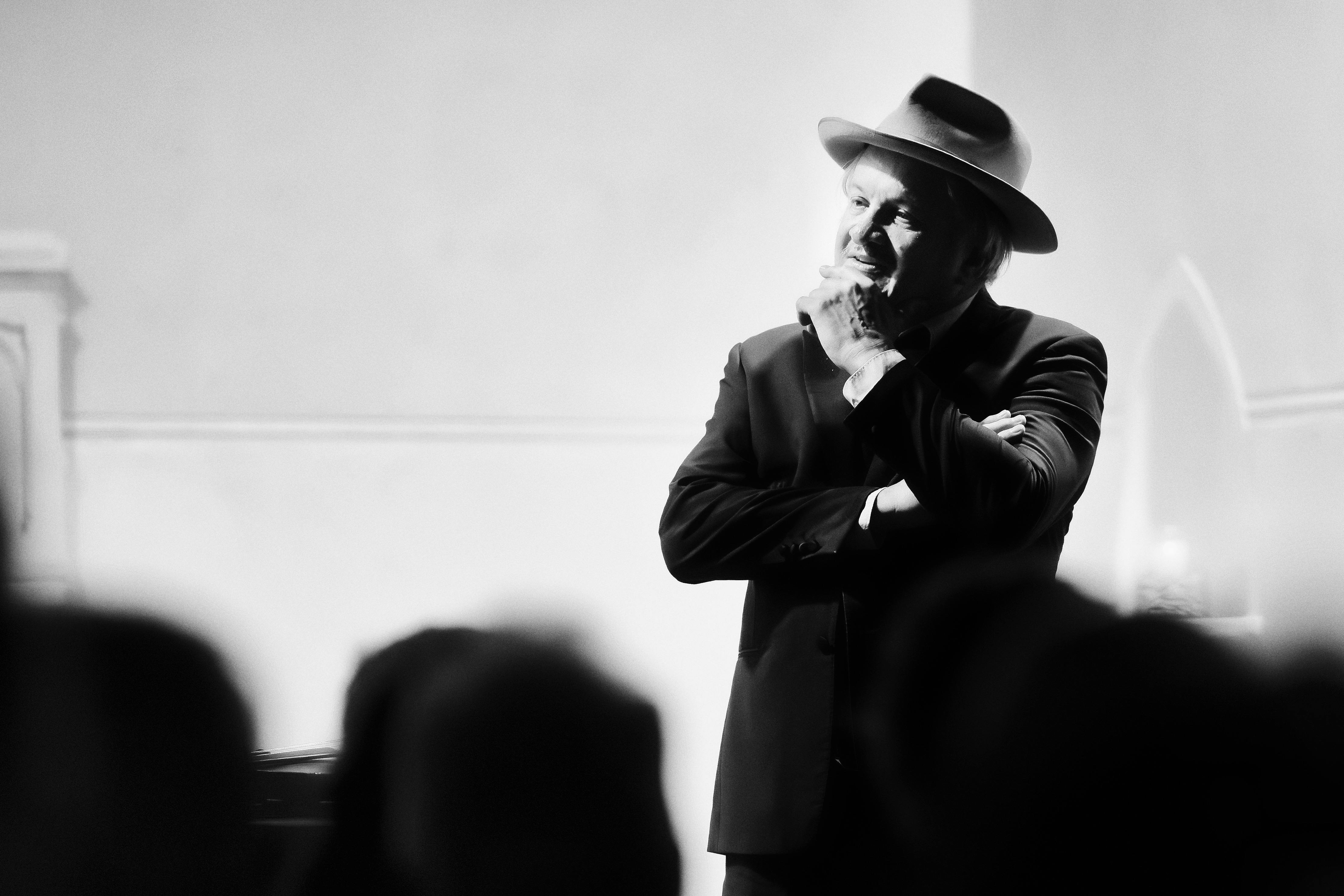 Black and white image of a man in a fedora with arms crossed and talking to an audience