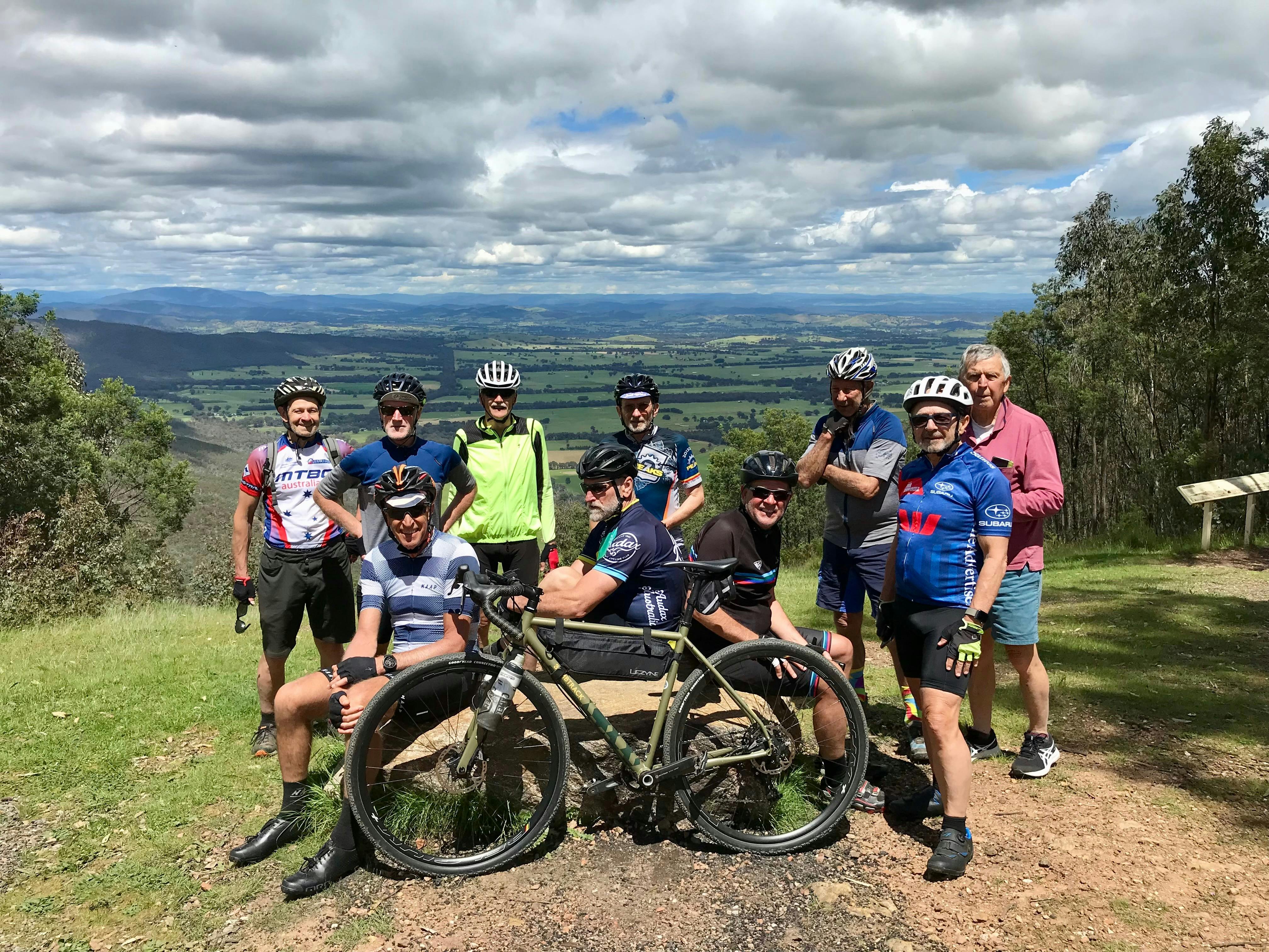 Group of cyclists resting at panic table at lookout with views of Murmungee Basin  and Mt Buffalo in