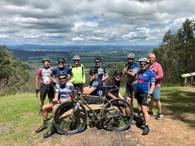 Group of cyclists resting at panic table at lookout with views of Murmungee Basin  and Mt Buffalo in