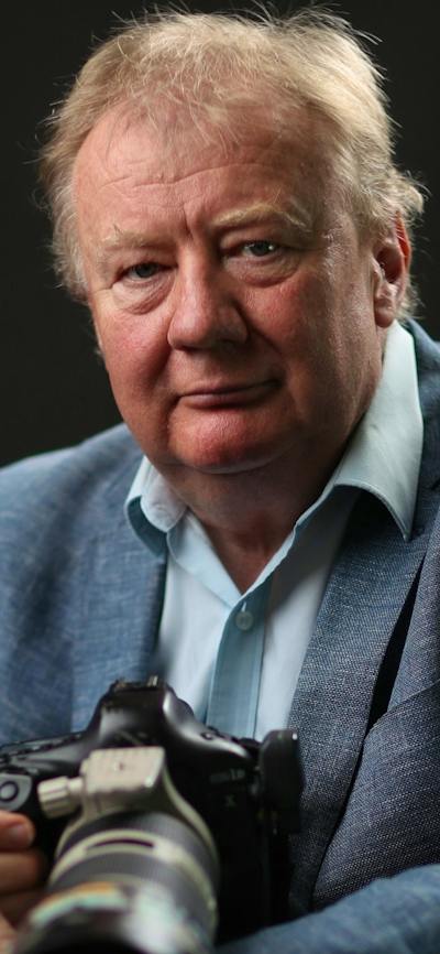A man with grey hair wearing a blue shirt and blazer, holding a large black camera.