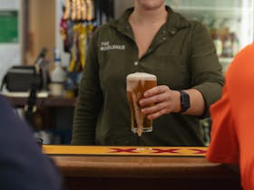 A front bar at a pub with a bar server holding out a frothy, overflowing beer to two patrons