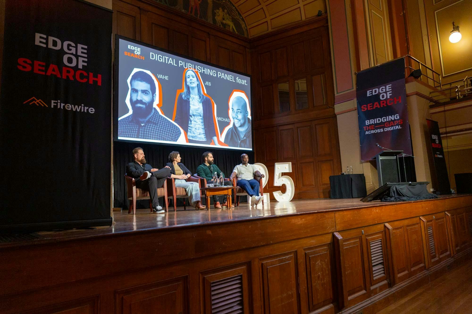 Four people sitting on stage in chairs in a panel style in front of a screen  with 3 people on it.