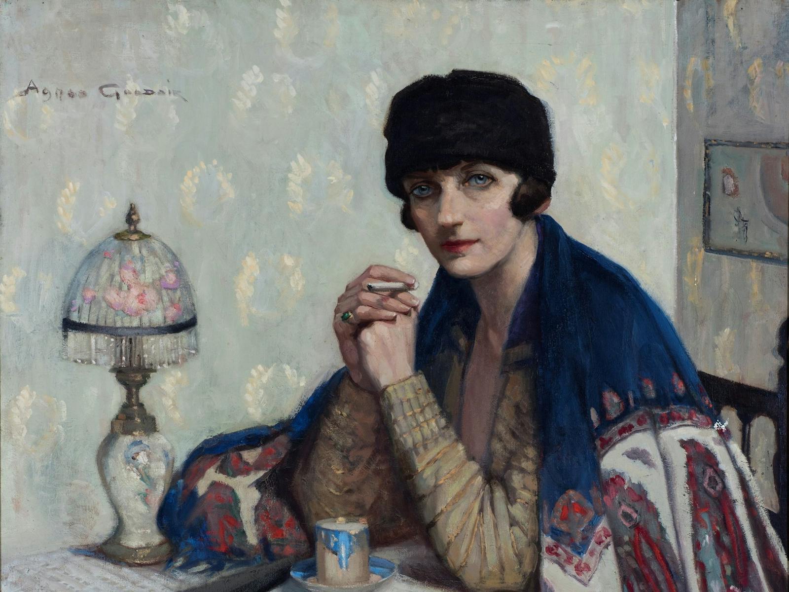 A woman in 1920s attire sits at a table with a patterned cloth. She wears a blue shawl and hat
