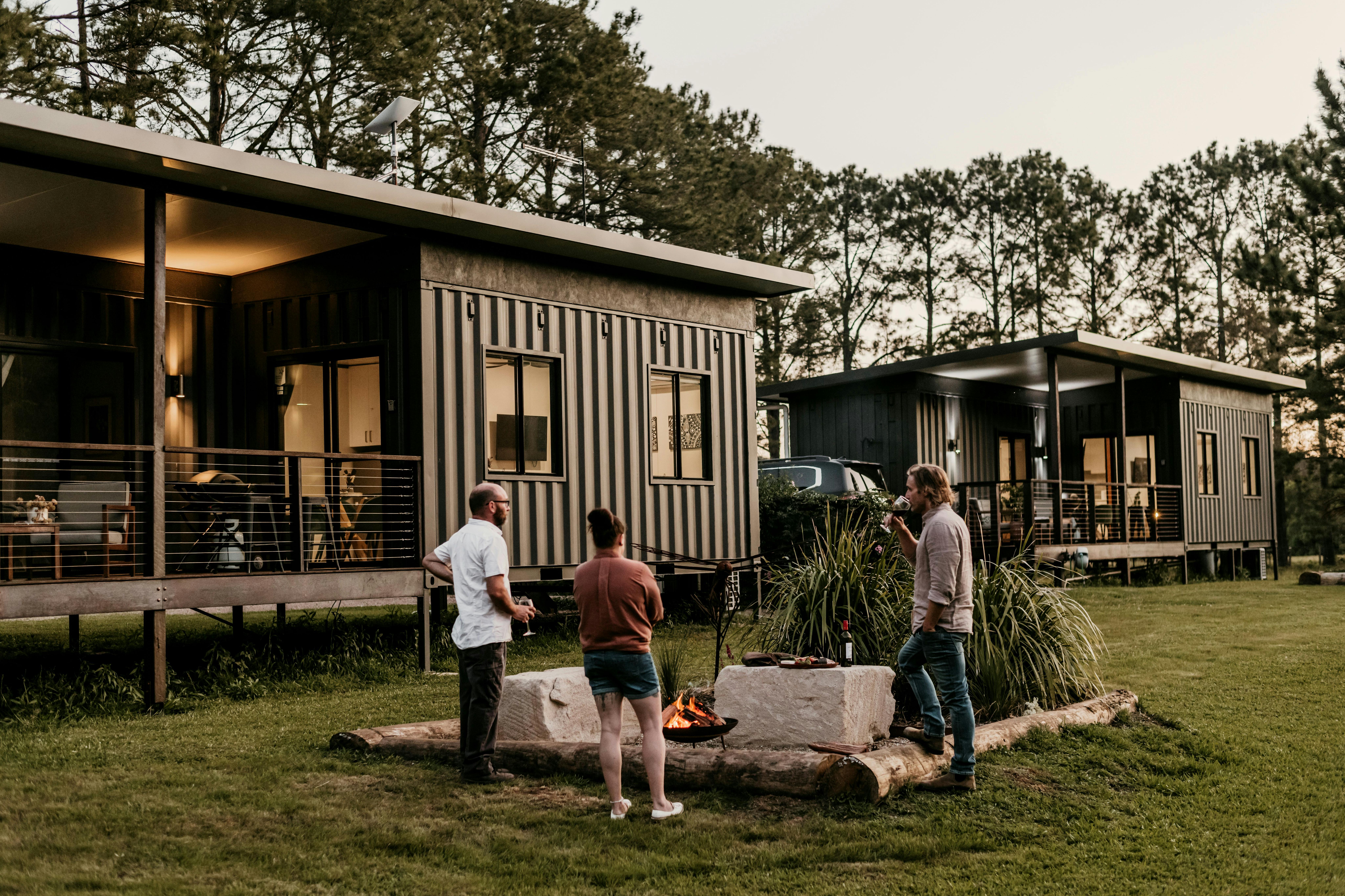 Three people enjoying wine by a firepit between two modern cabins made from shipping containers.