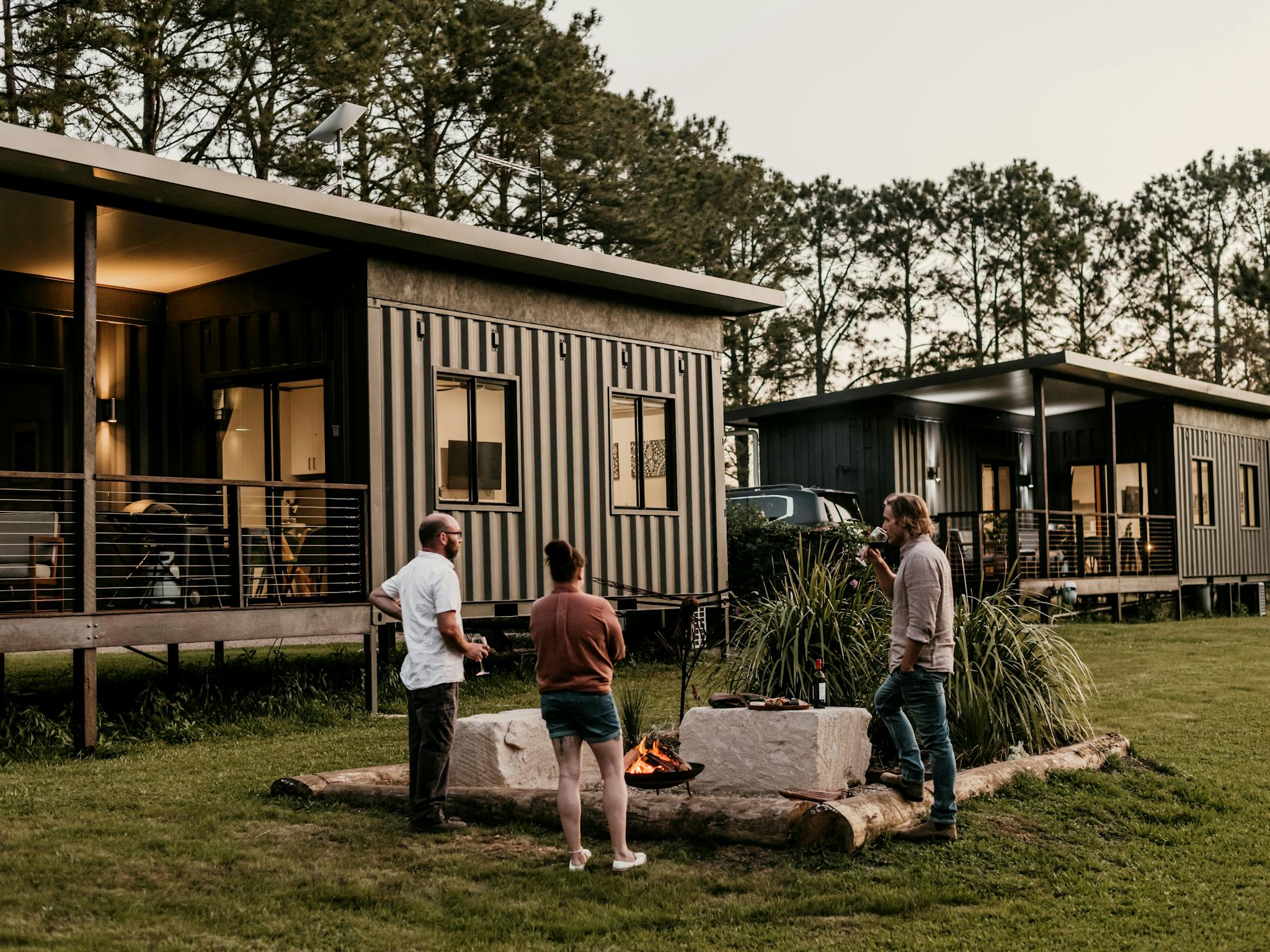 Three people enjoying wine by a firepit between two modern cabins made from shipping containers.