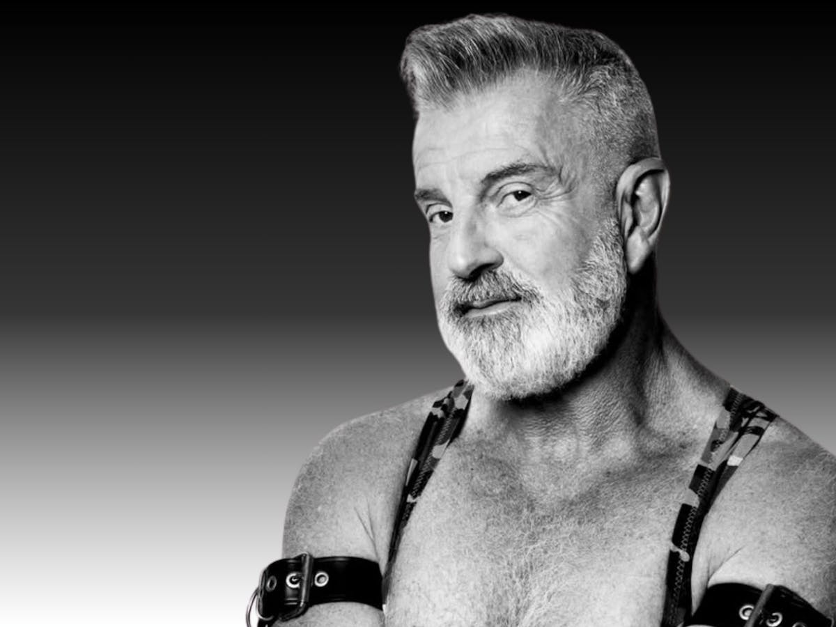 A mature man with grey hair wearing a leather chest harness.. Black and white portrait.