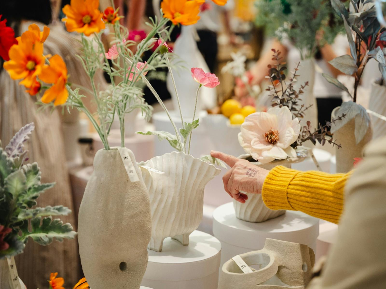 White ceramic works on display with colourful flowers and a patron pointing with a yellow sleeve