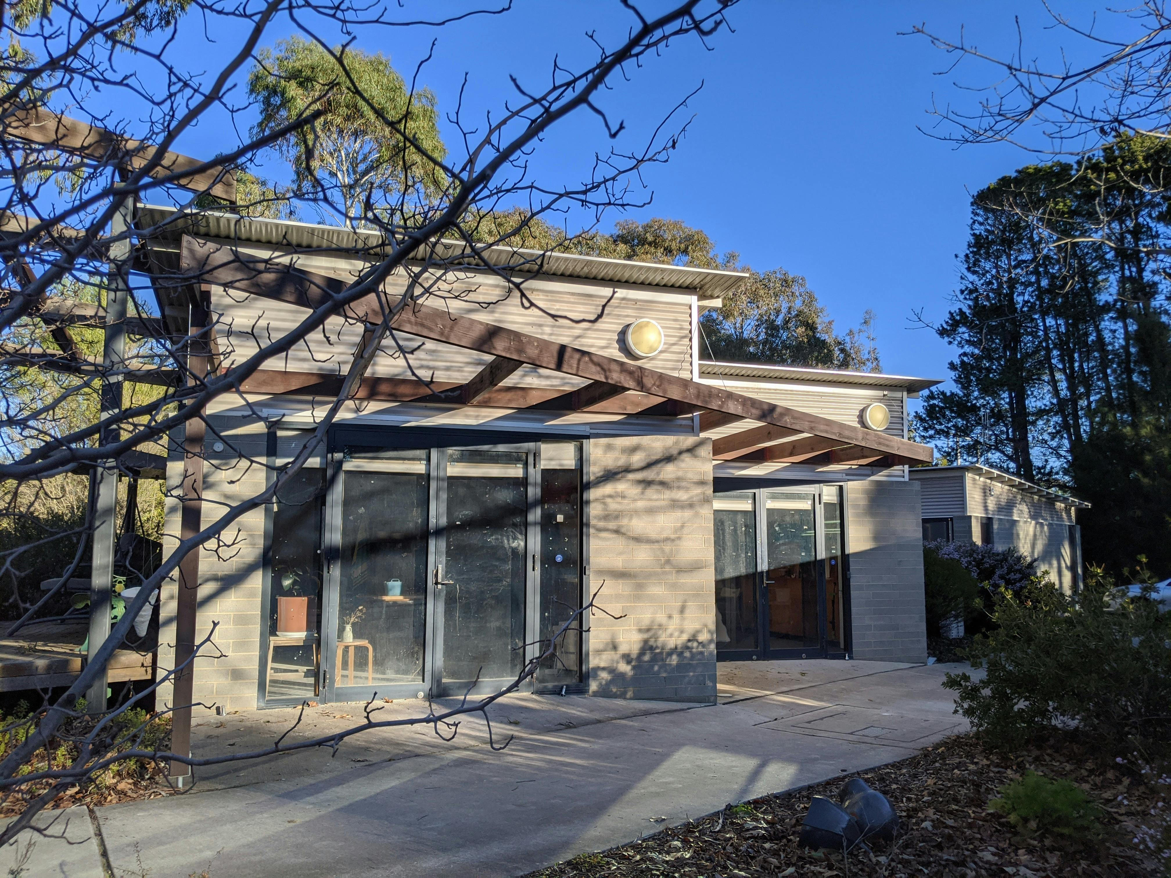 A small grey concrete building with a brown pergola structure out the front.  Clear blue skies.