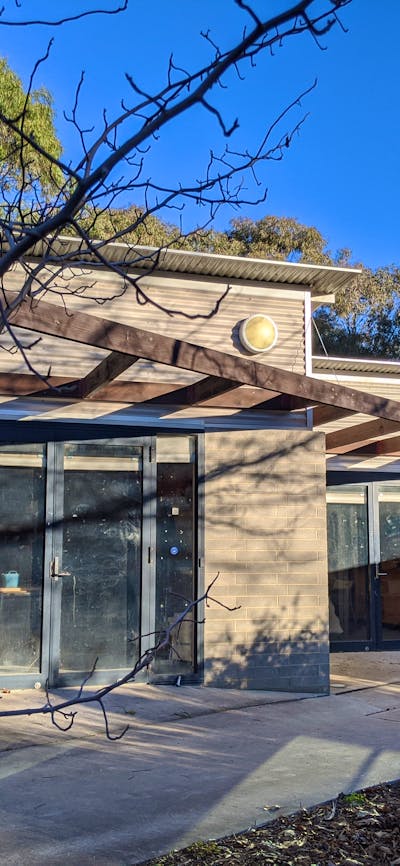 A small grey concrete building with a brown pergola structure out the front. Clear blue skies.