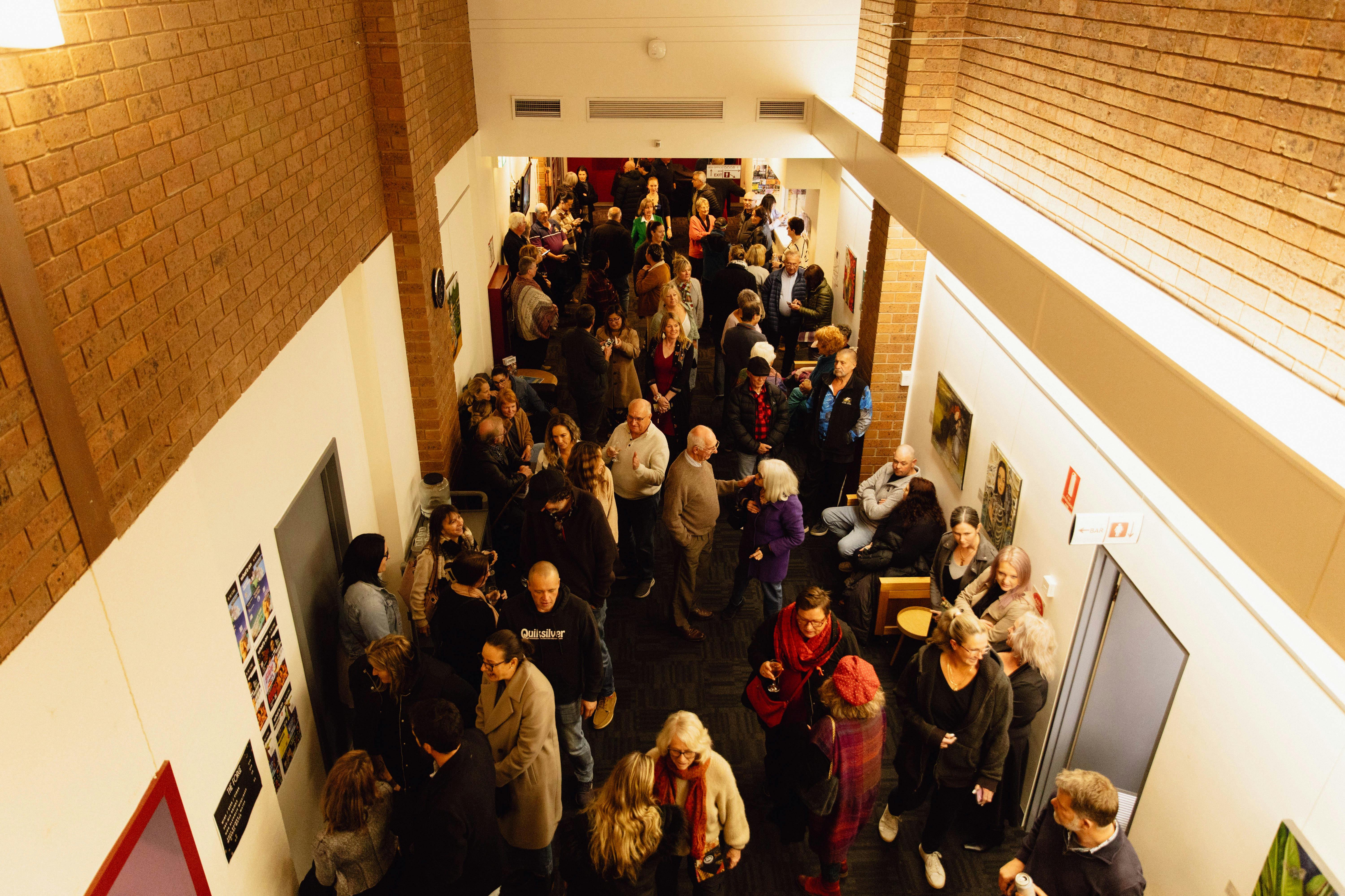 Lots of people are gathered together in a narrow passage way which is the foyer of the venue