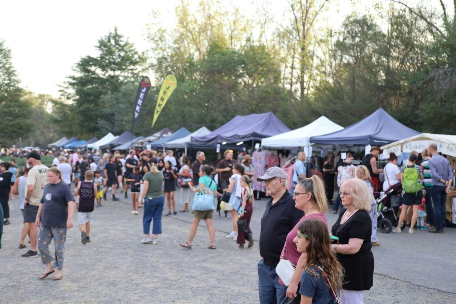 people walking around an open ground with market stalls with the trees in the background