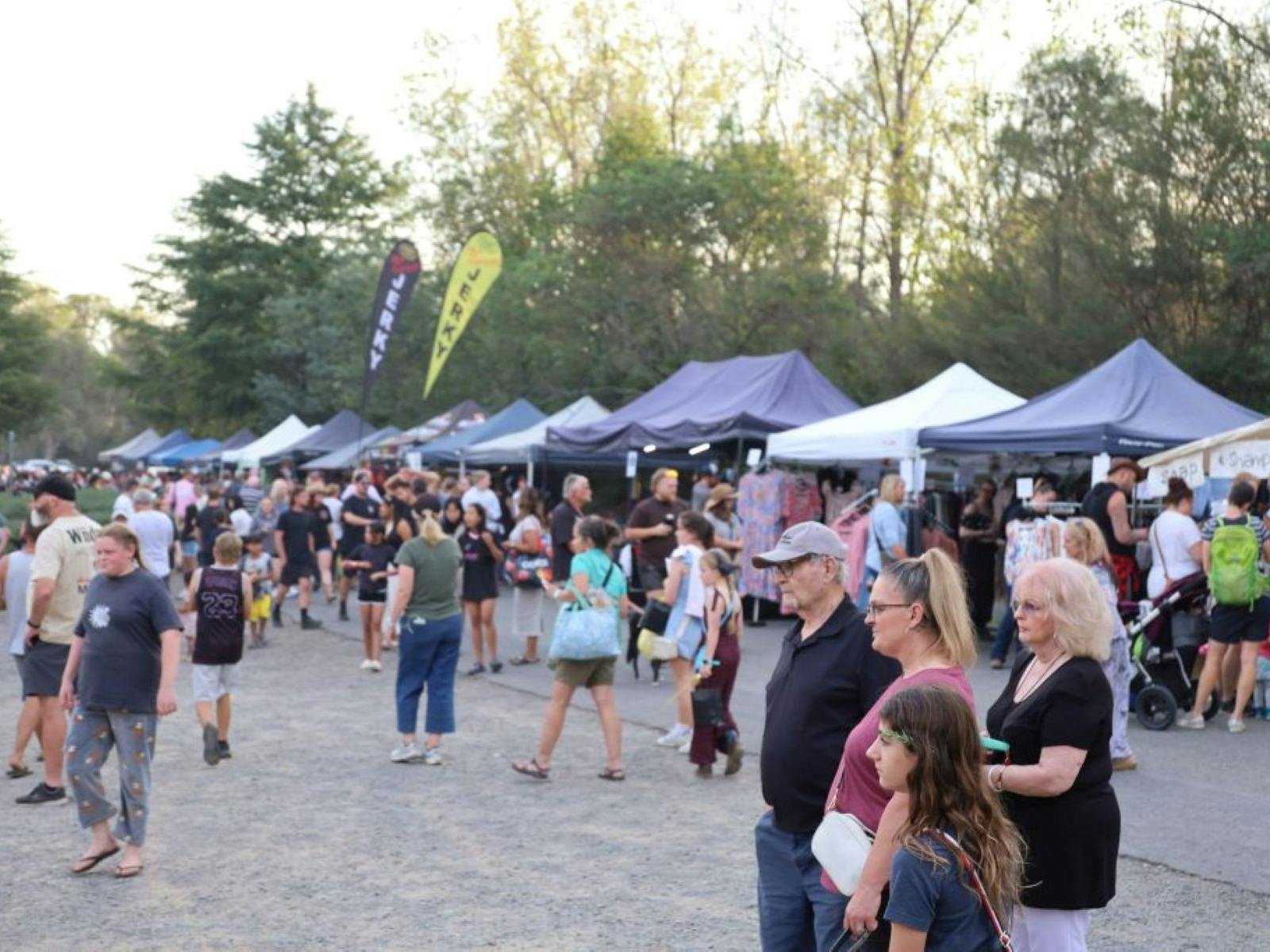 people walking around an open ground with market stalls with the trees in the background