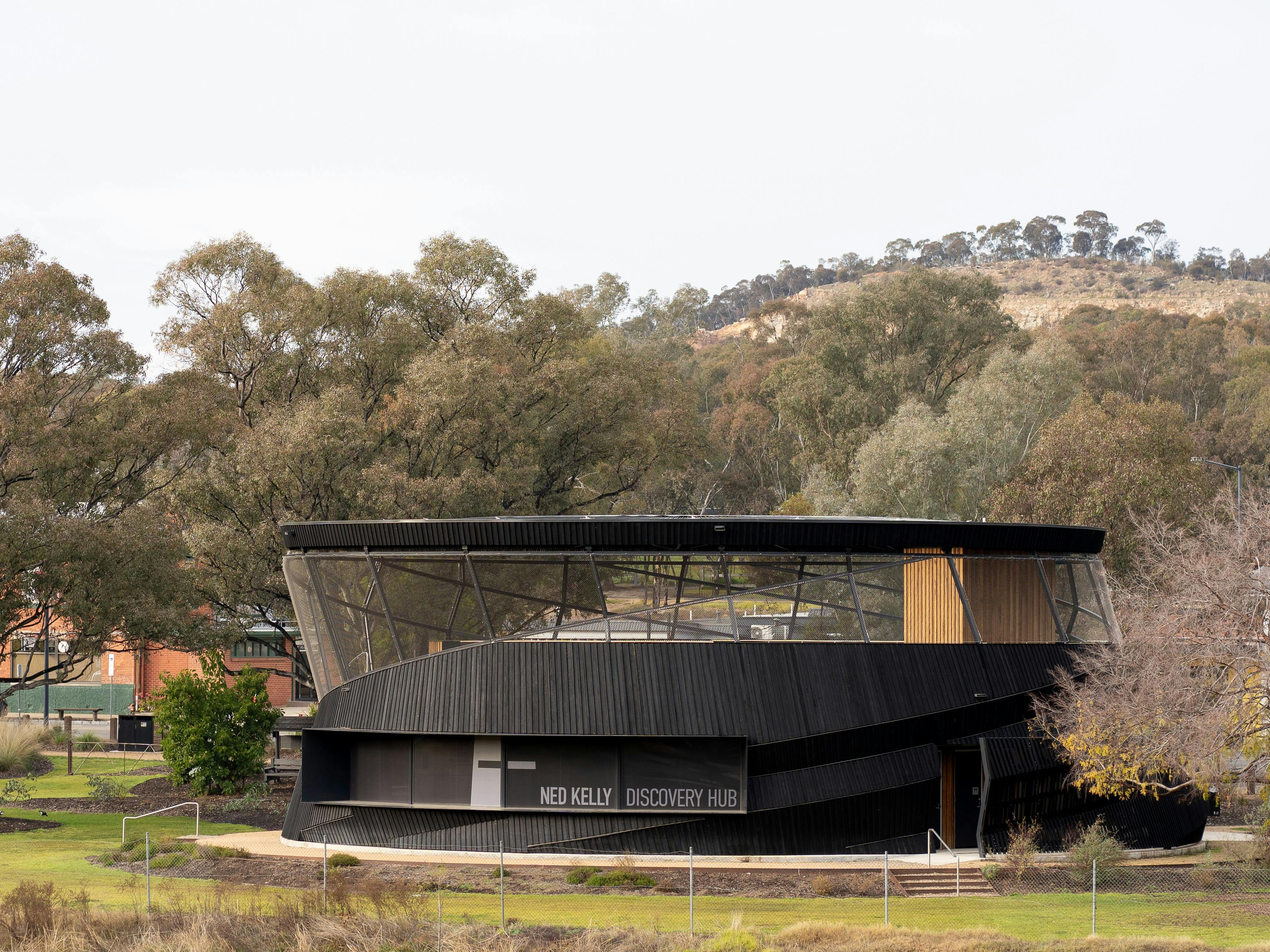 Modern, curved Ned Kelly Discovery Hub building set among trees with a hillside backdrop Glenrowan