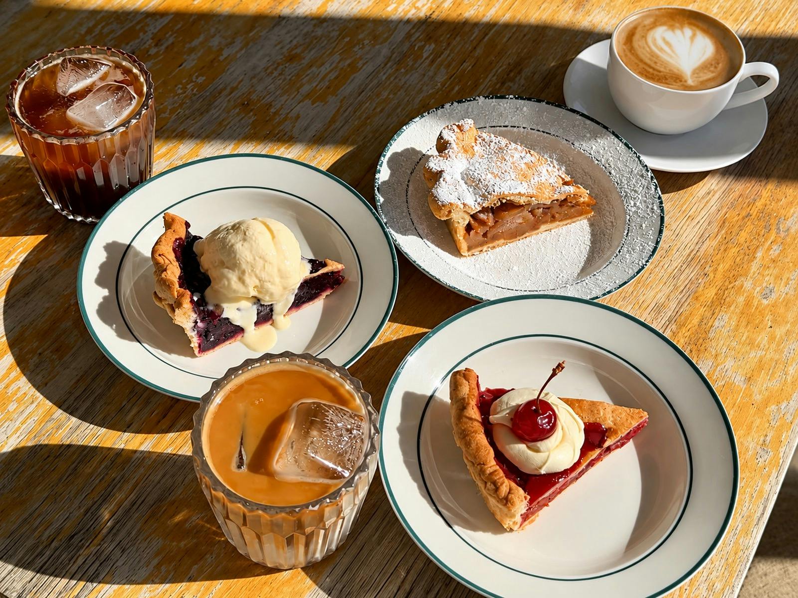 A sunlit table with slices of cherry, blueberry and apple pie served alongside iced and hot coffees.