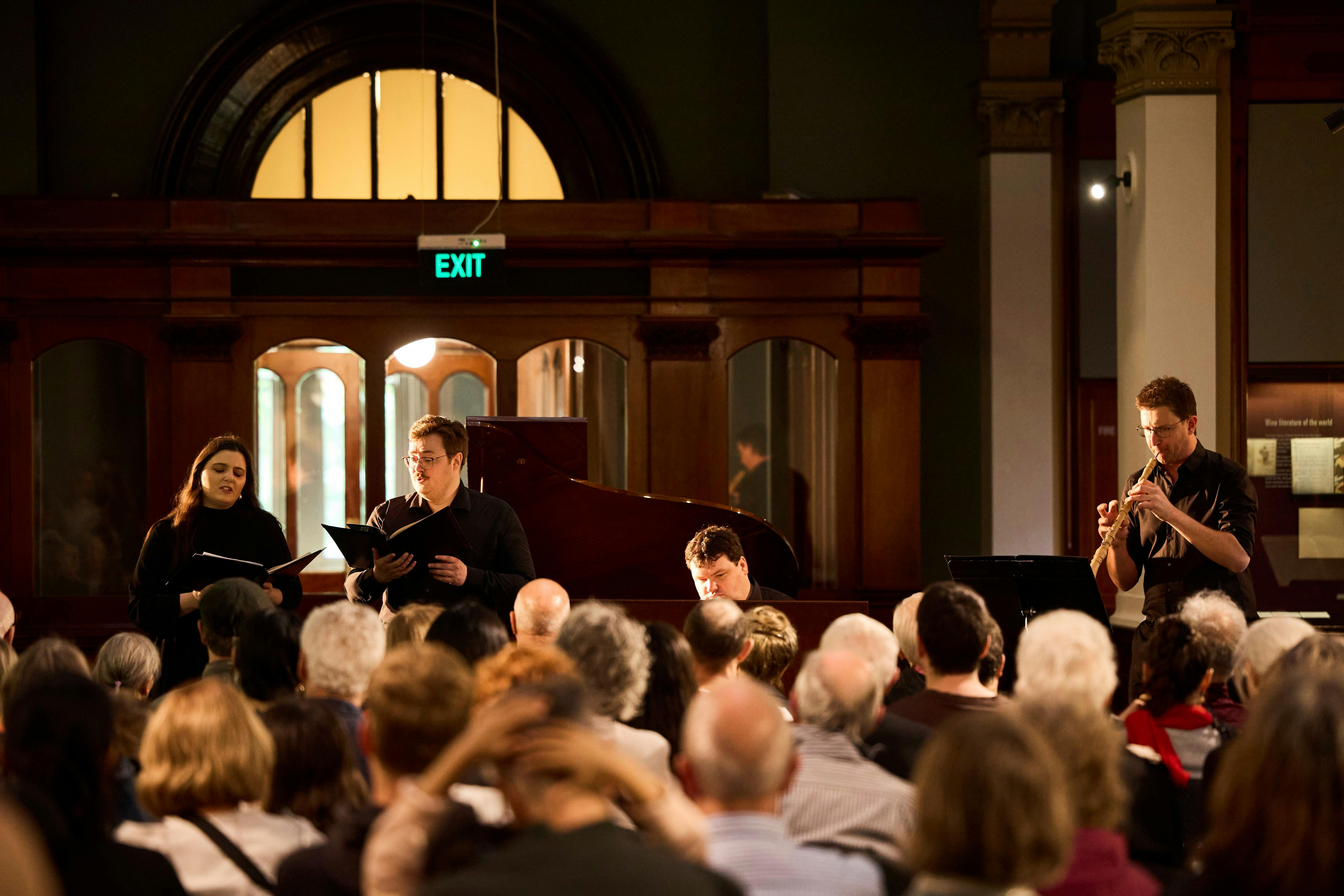 A seated audience watch a musical performance by two singers, accompanied by a pianist and oboist.