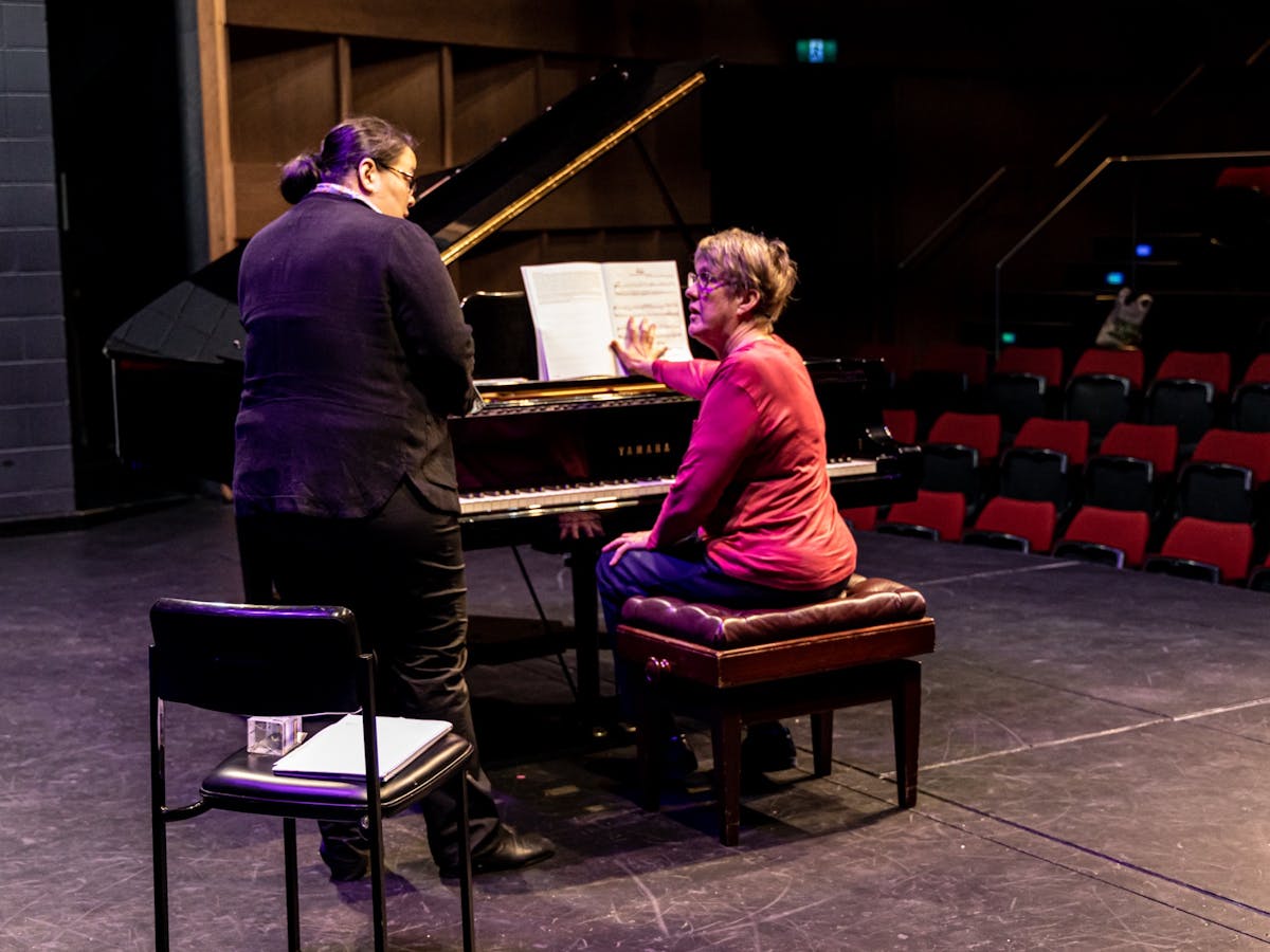 A student, seated the piano, receives guidance from the teacher
