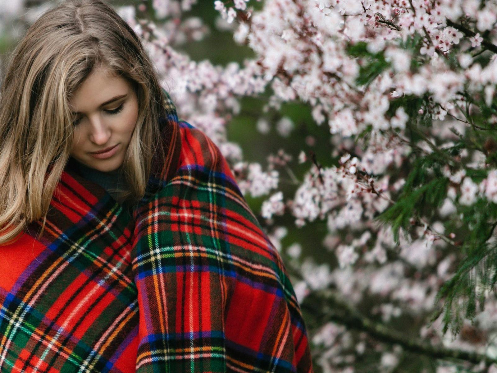 Young lady with long brown hair wrapped in red, green, blue checked blanket in front of pink blossom