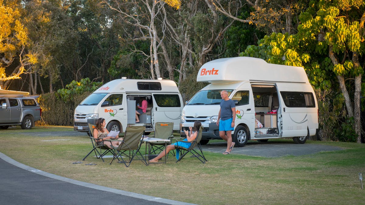 Image of 2 Britz campervans on site with people in in camp chairs in the late afternoon sun