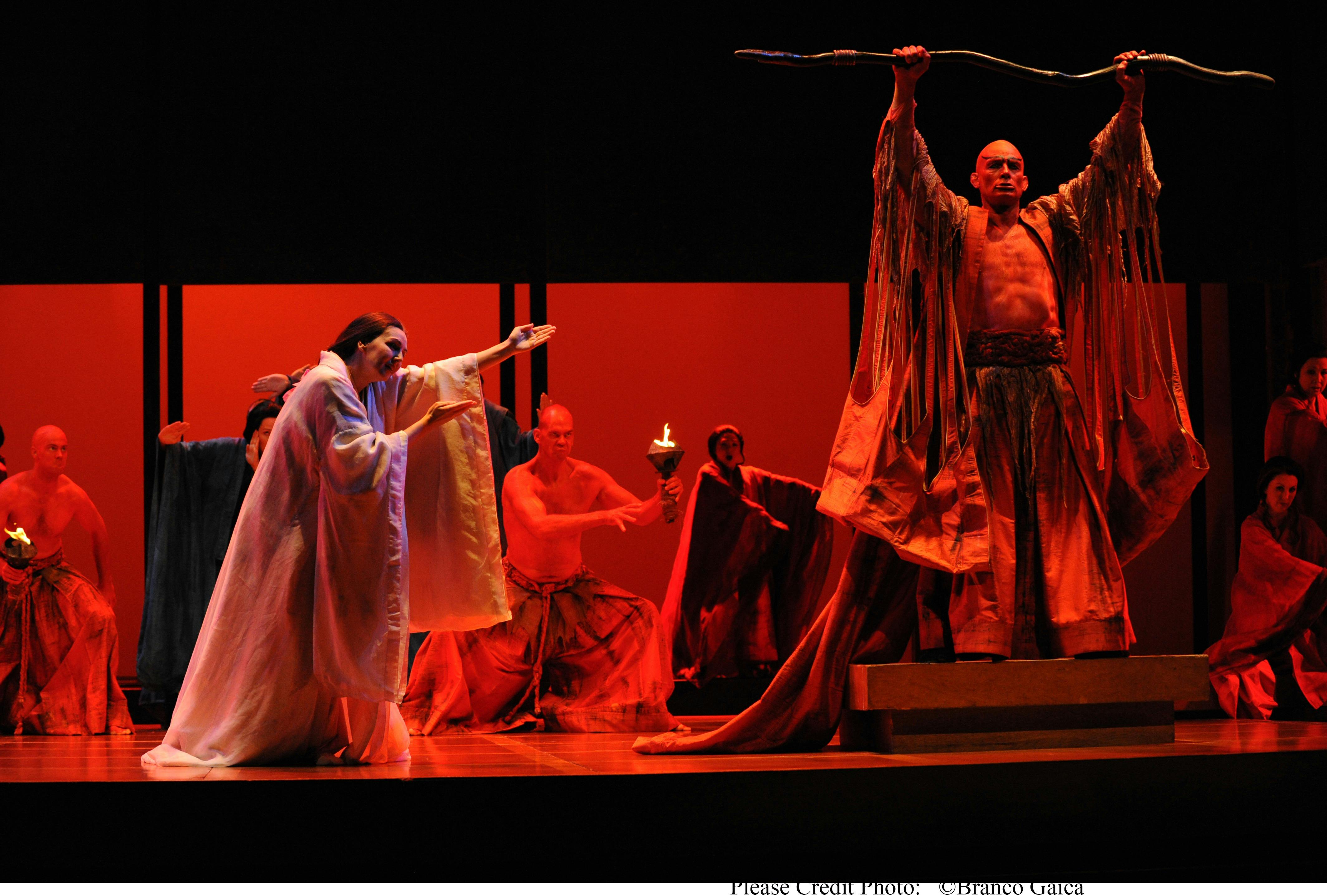 On a red-lit stage, a woman  as a reaches her arms out as a robed man raises a staff.