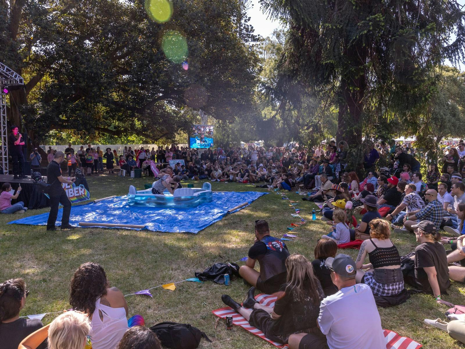People sit around an inflatable pool where two people are standing inside, wrestling.