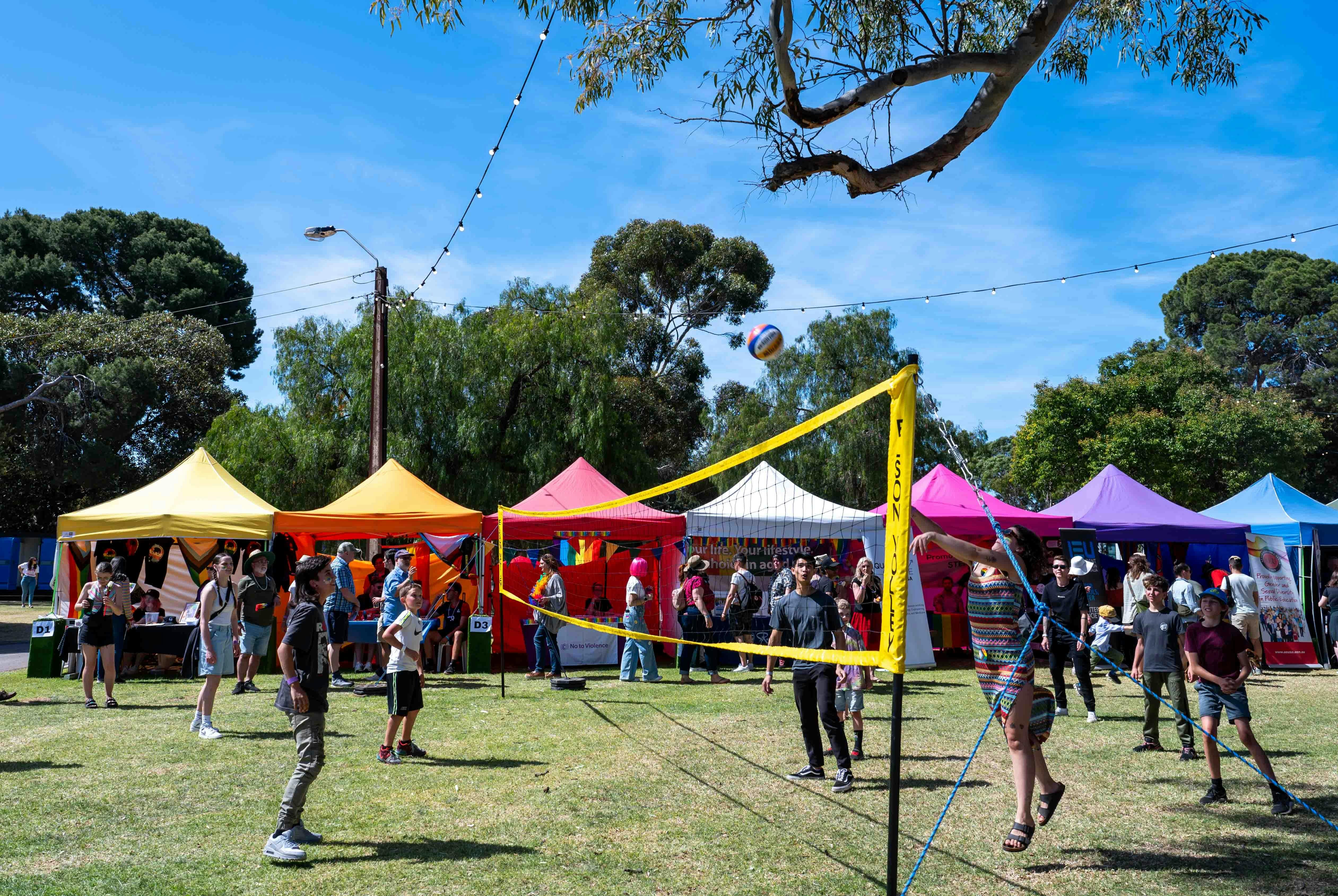 People playing volleyball in a park with rainbow-coloured gazebo stalls in the background.