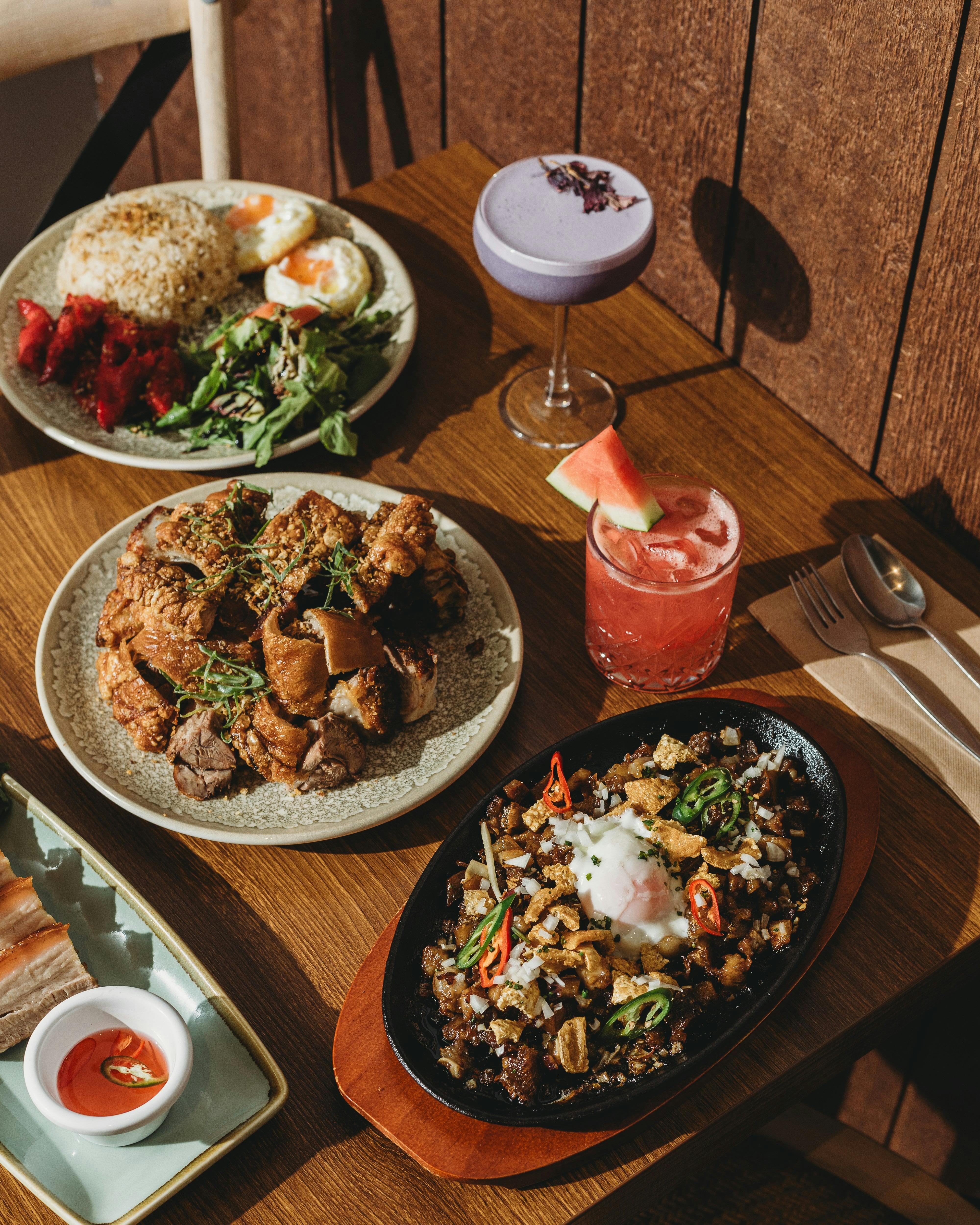 A wooden table filled with Filipino dishes including sizzling sisig with egg, crispy pork pieces, a