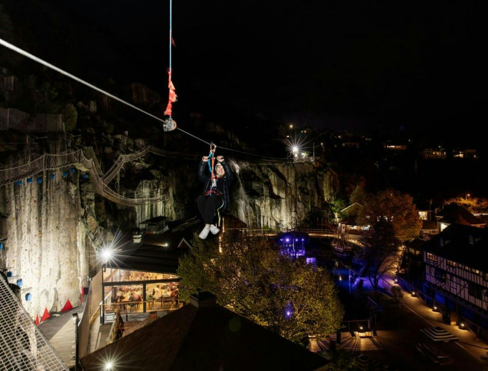 Participant Ziplining at nighttime. The park complex and natural cliff face beautifully lit
