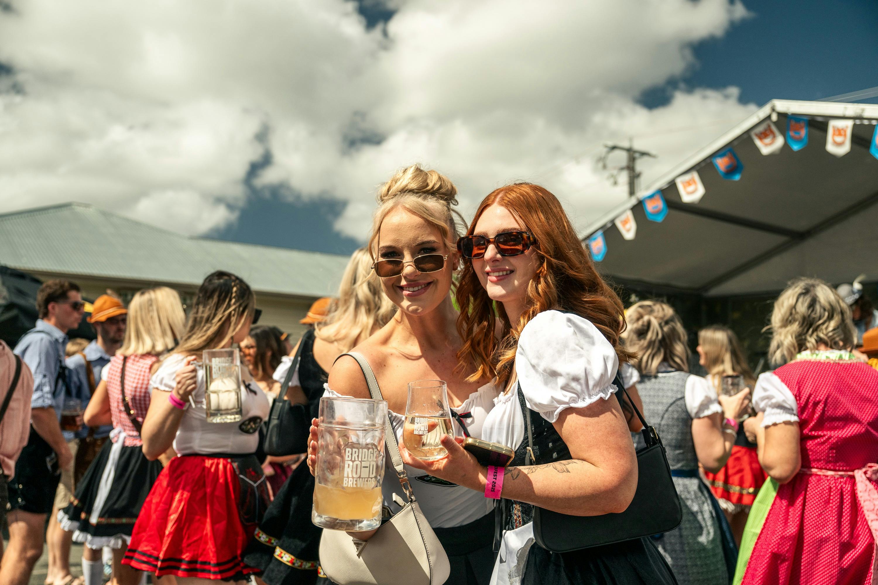 Two women smiling outside surrounded by crowd giving cheers with their beers.