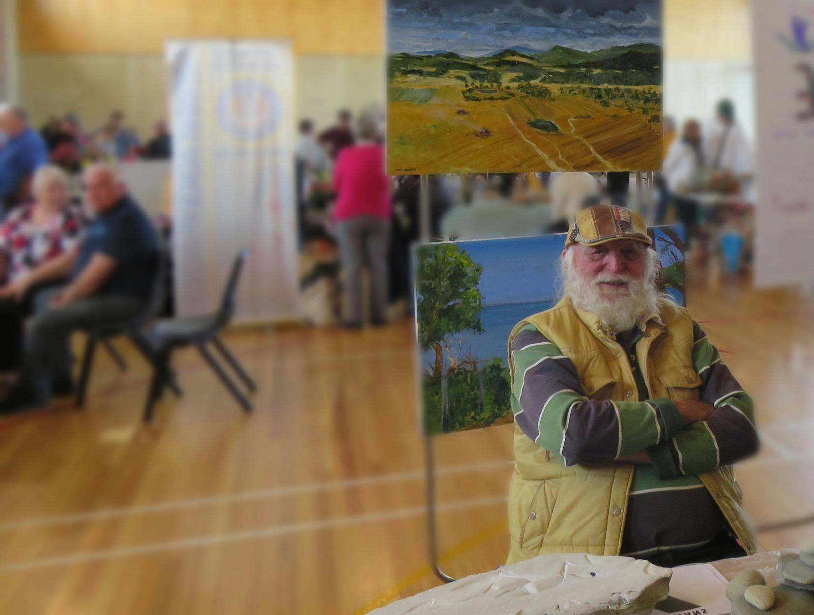 A white bearded man sits in front of some landsacpe paintings
