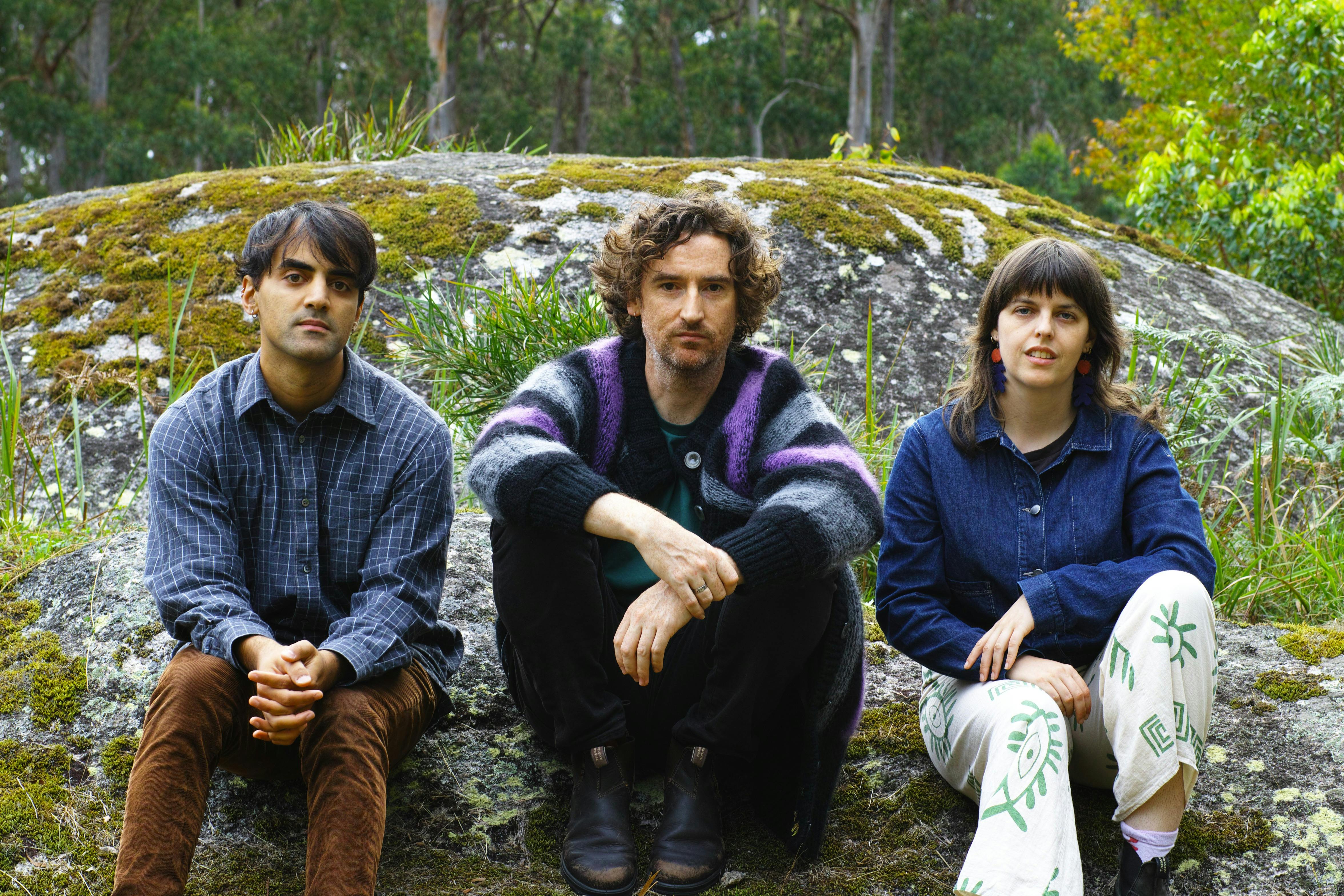 Image of three people sitting on a rock in the bush with a large mossy boulder behind them.