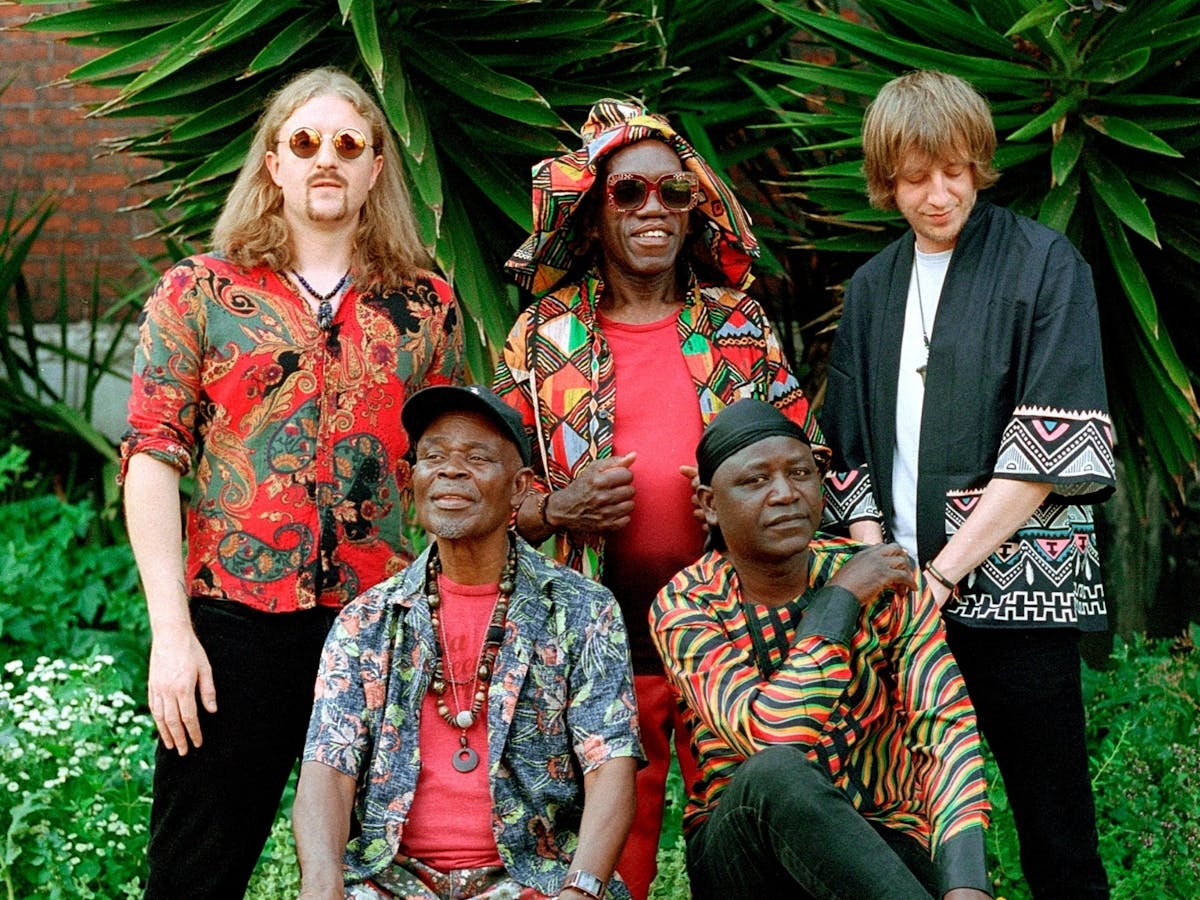 A group of five men pose together outdoors in front of lush green plants. The group appears cheerful