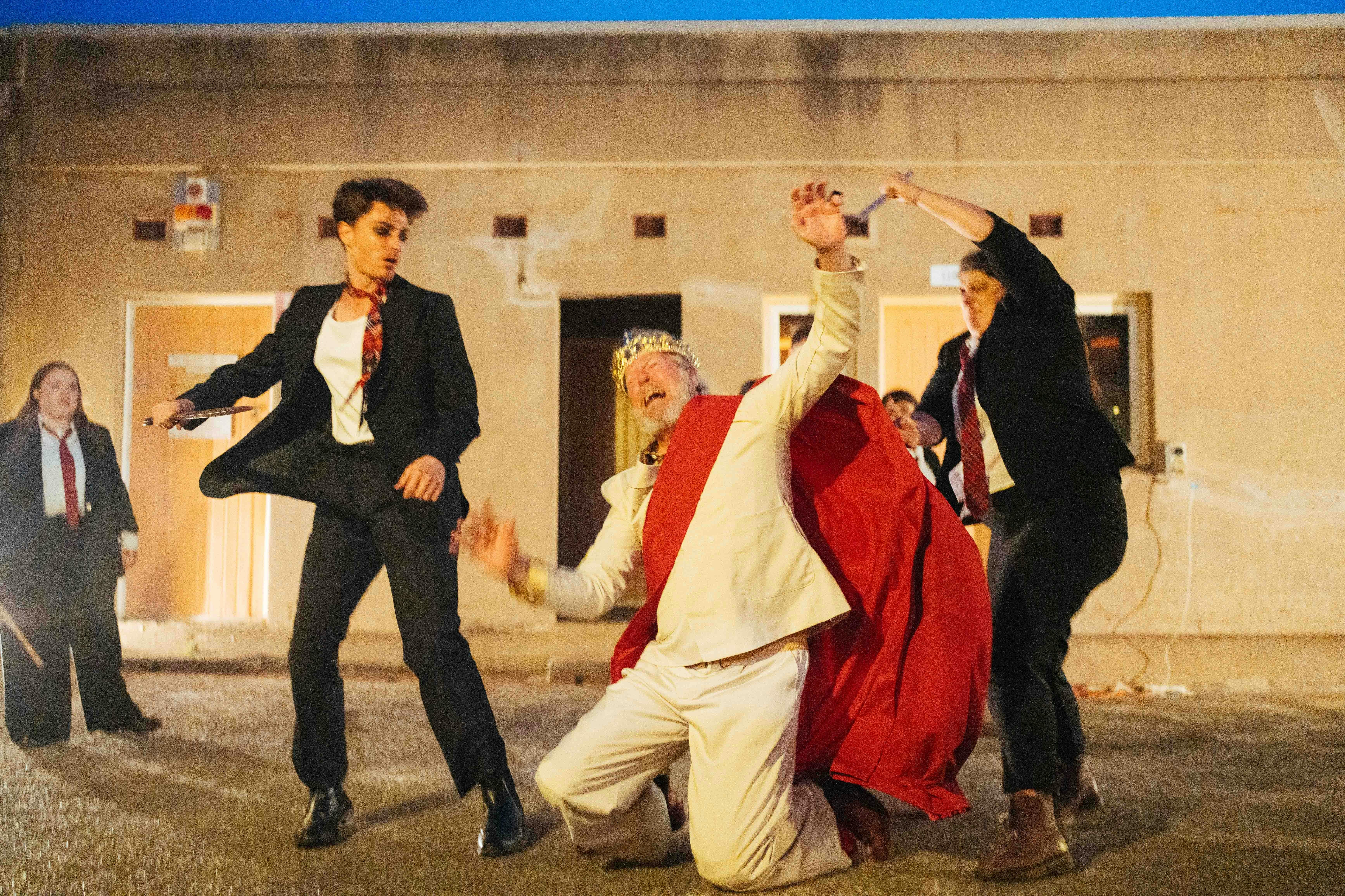 Two suited actors with fake swords strike at an older man dressed like a King—he falls to the ground