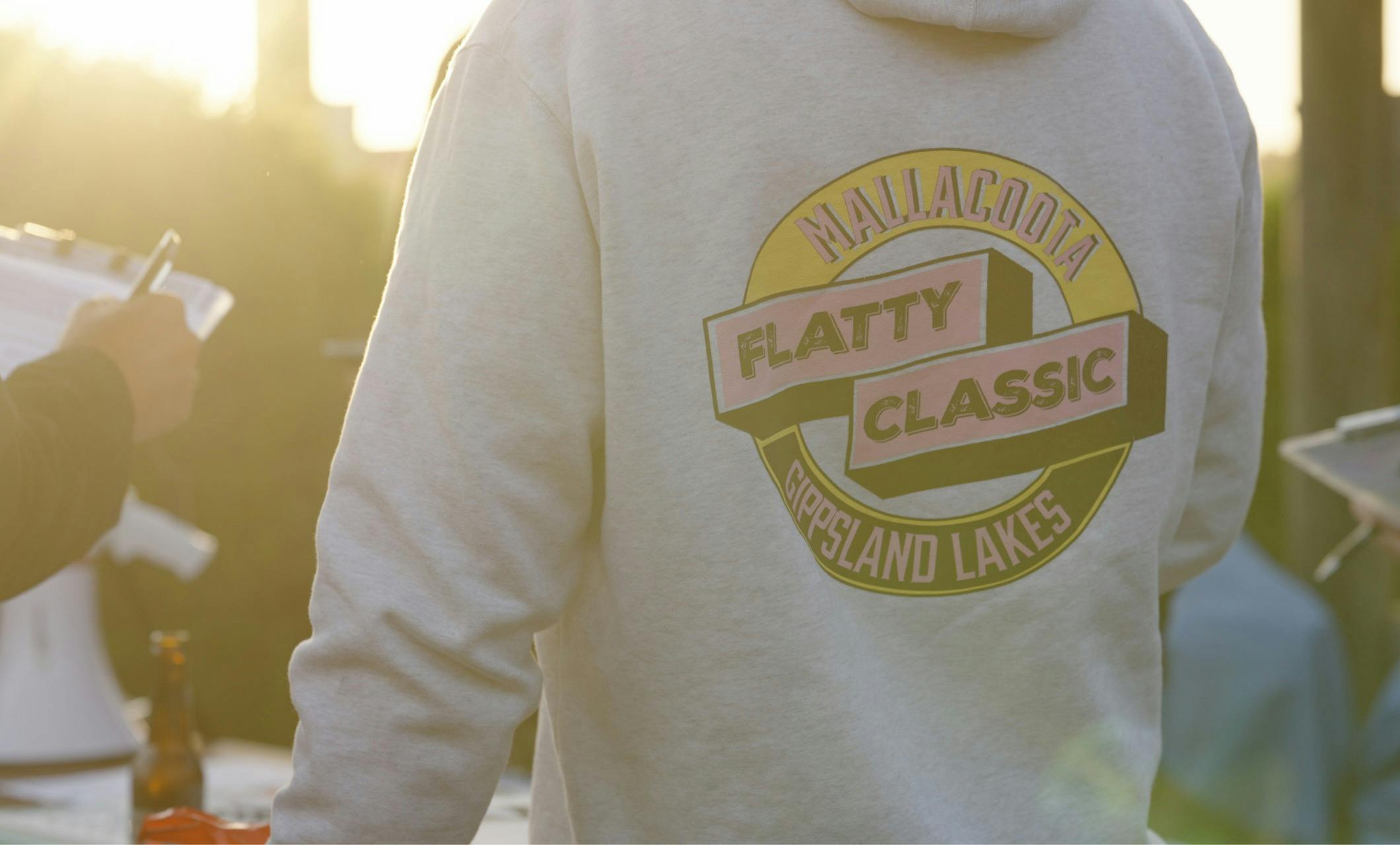 Back of a cream hoodie showing the Mallacoota Flatty Classic logo, golden hour light behind