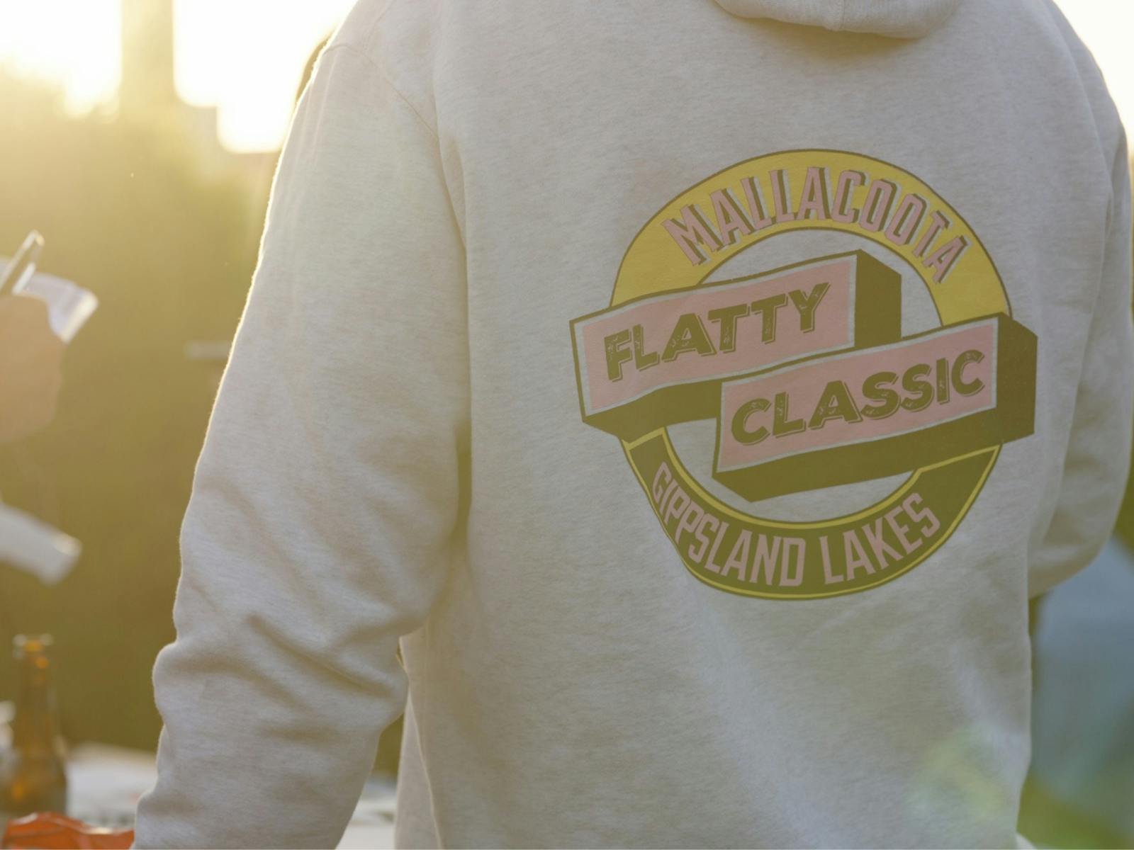 Back of a cream hoodie showing the Mallacoota Flatty Classic logo, golden hour light behind